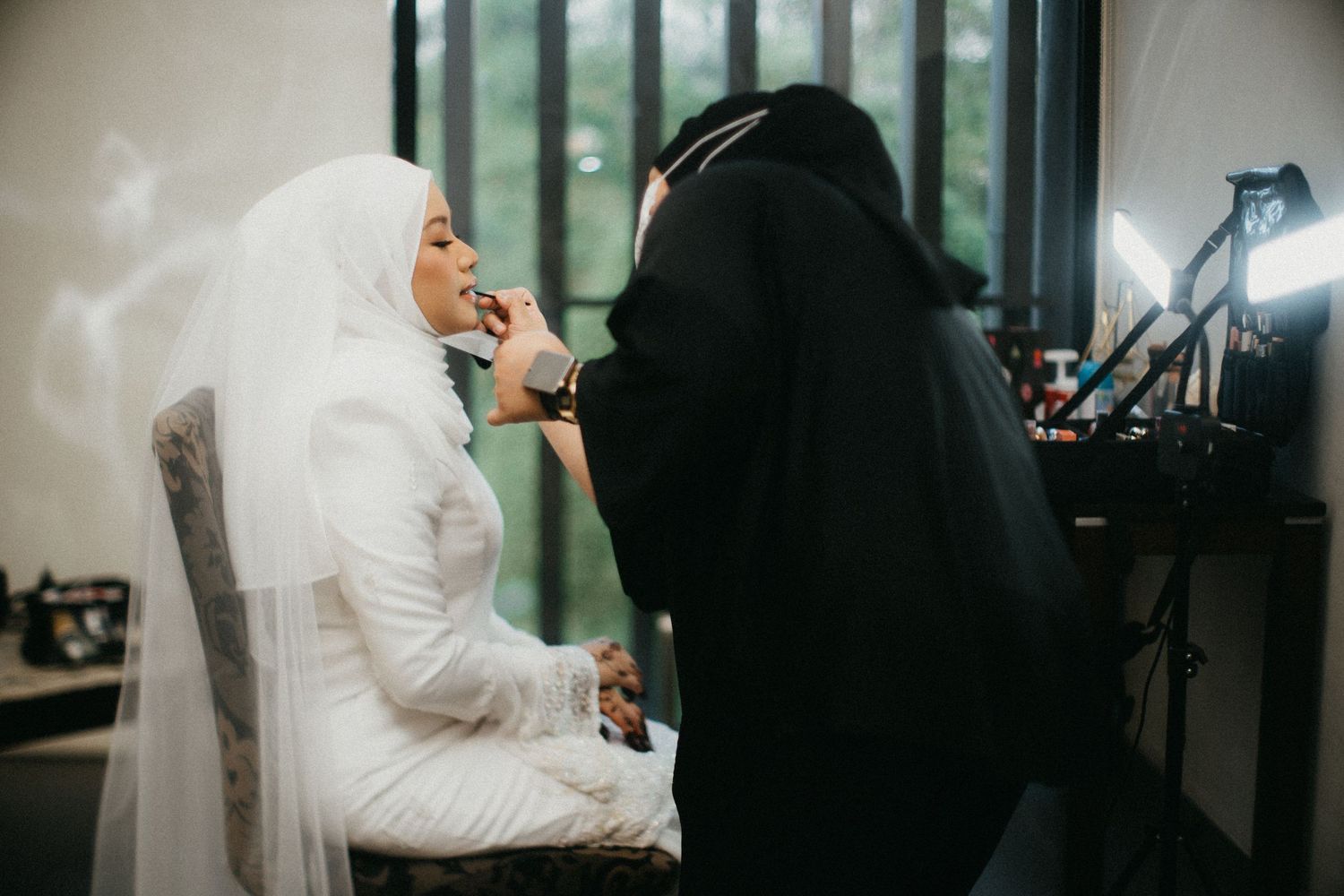 A makeup artist applies cosmetics to someone wearing a white hijab and outfit during a beauty preparation session.