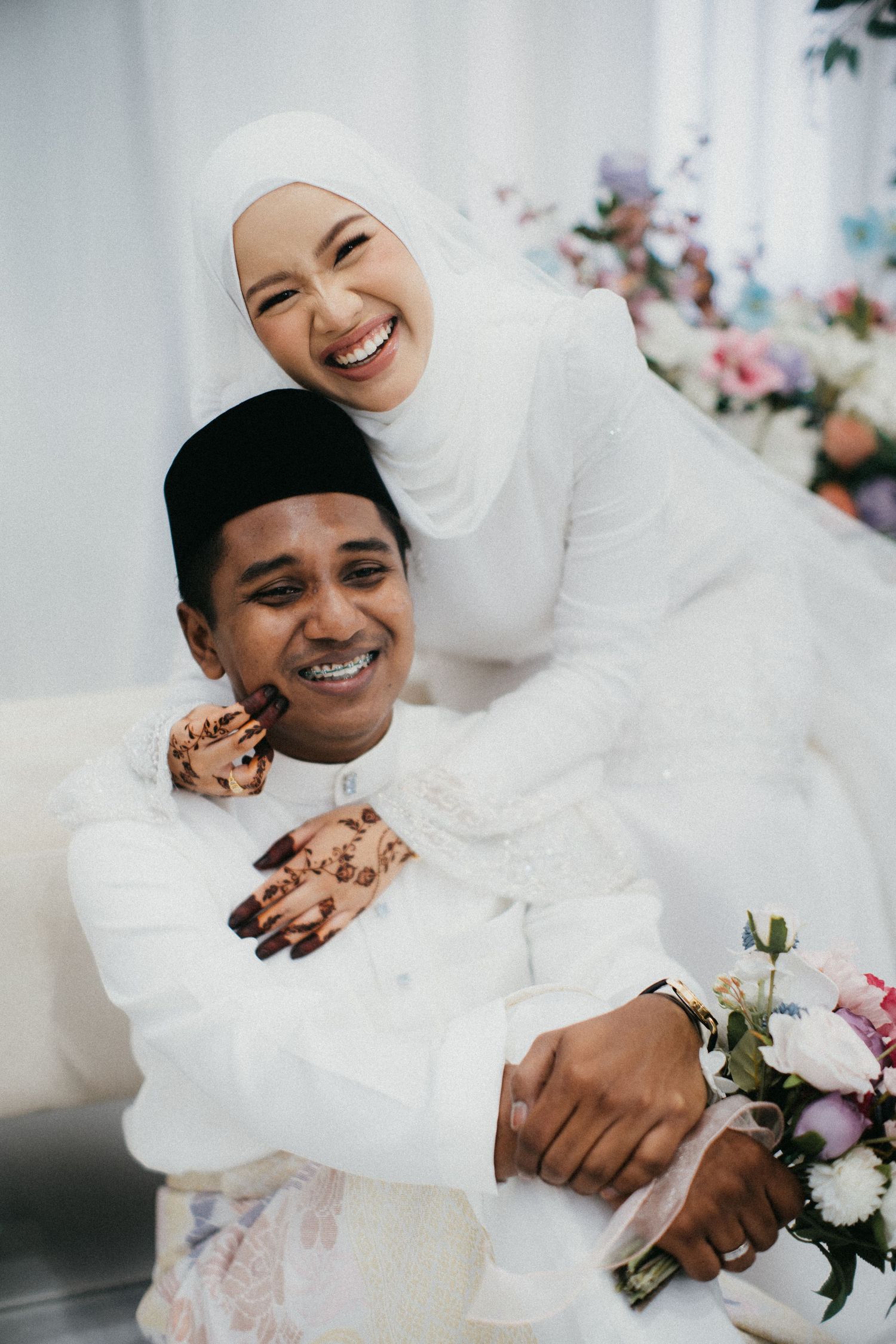 A joyful wedding couple in matching white traditional attire embrace while smiling during their celebration.