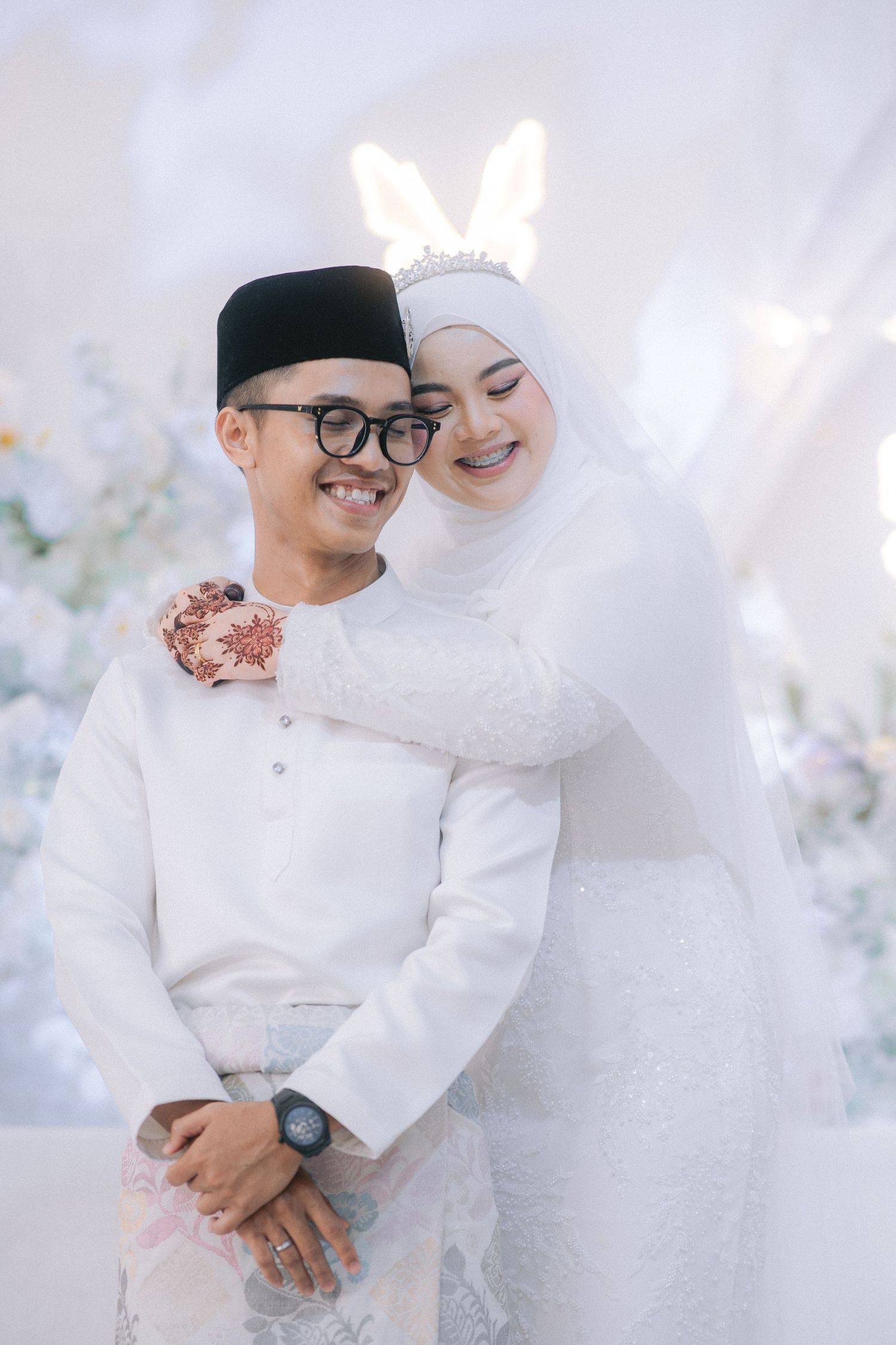 Two people in traditional white wedding attire embrace during a romantic photoshoot with light floral decor.