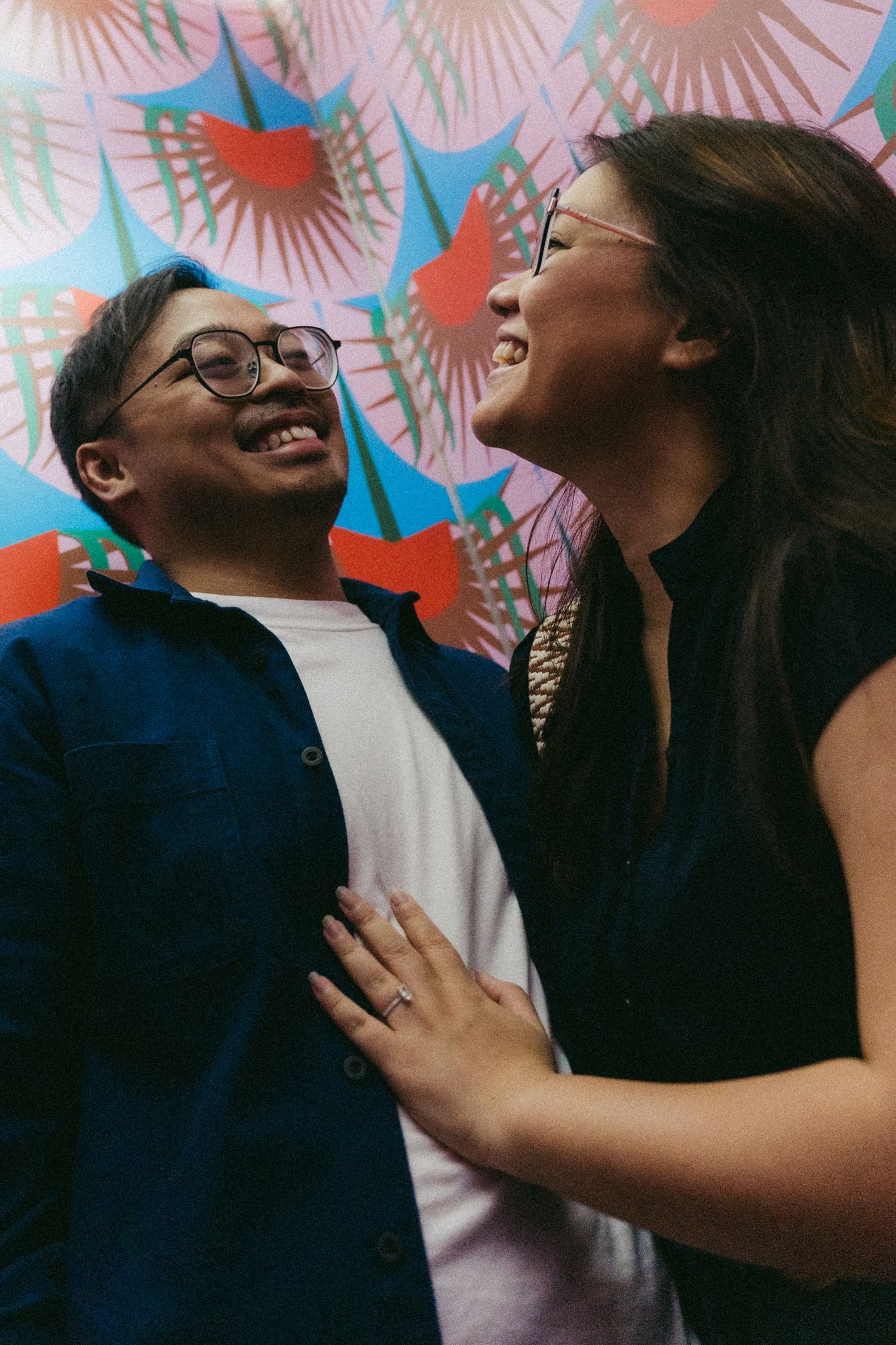 A joyful couple sharing a candid laugh together against a vibrant background with pink and blue fan patterns.