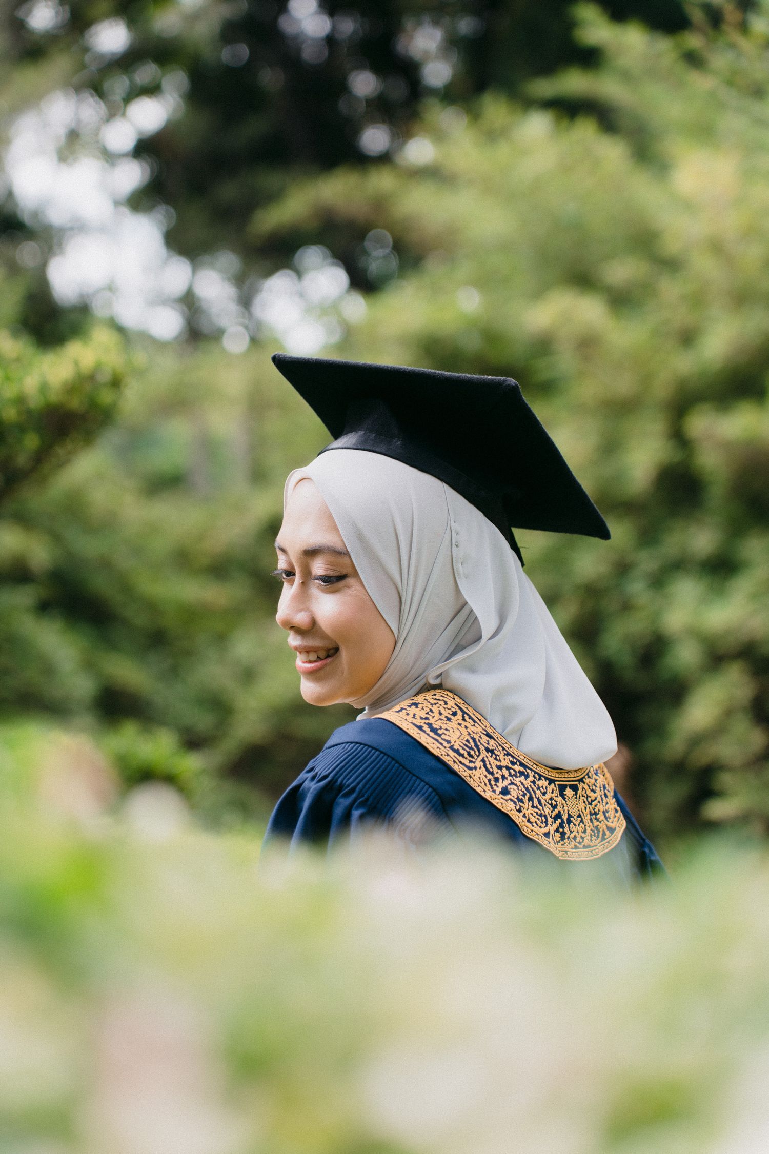 A graduate in a navy blue gown and white hijab with ornate gold trim smiles while wearing a mortarboard cap outdoors.