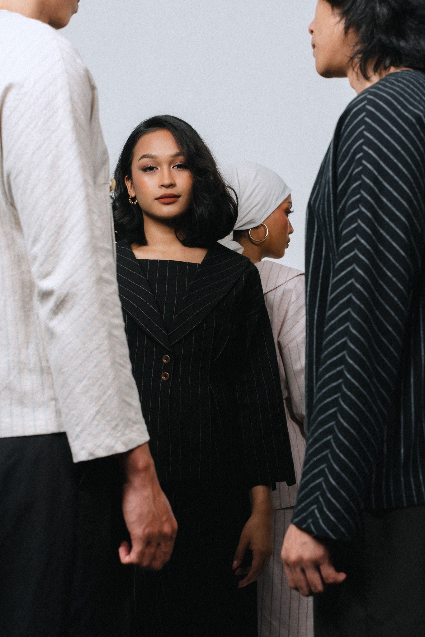 A young woman in a black pinstripe blazer stands among a group of people in neutral toned outfits against a gray background.