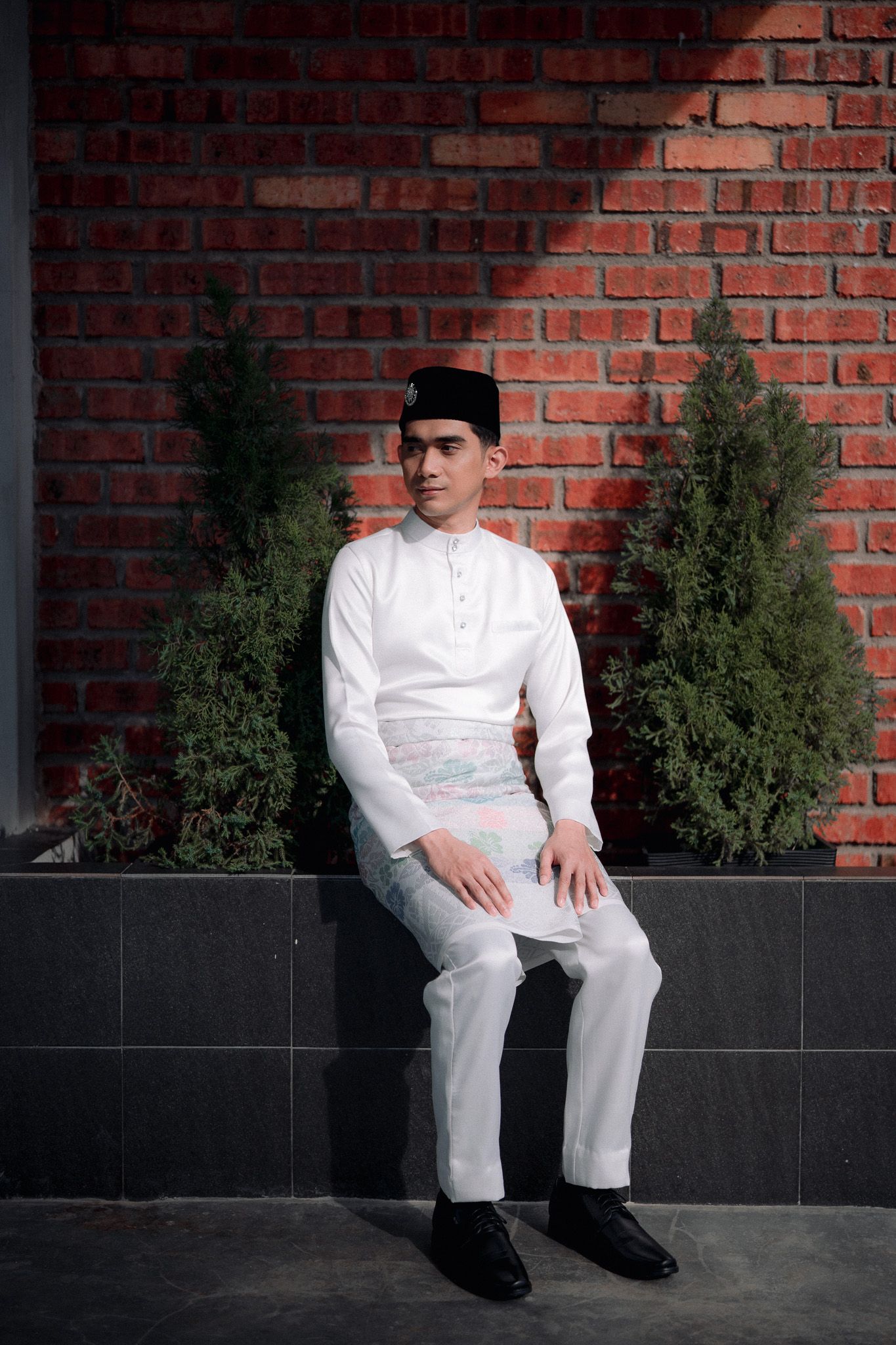 Young man in white baju melayu and songkok sitting against a red brick wall with green shrubs.