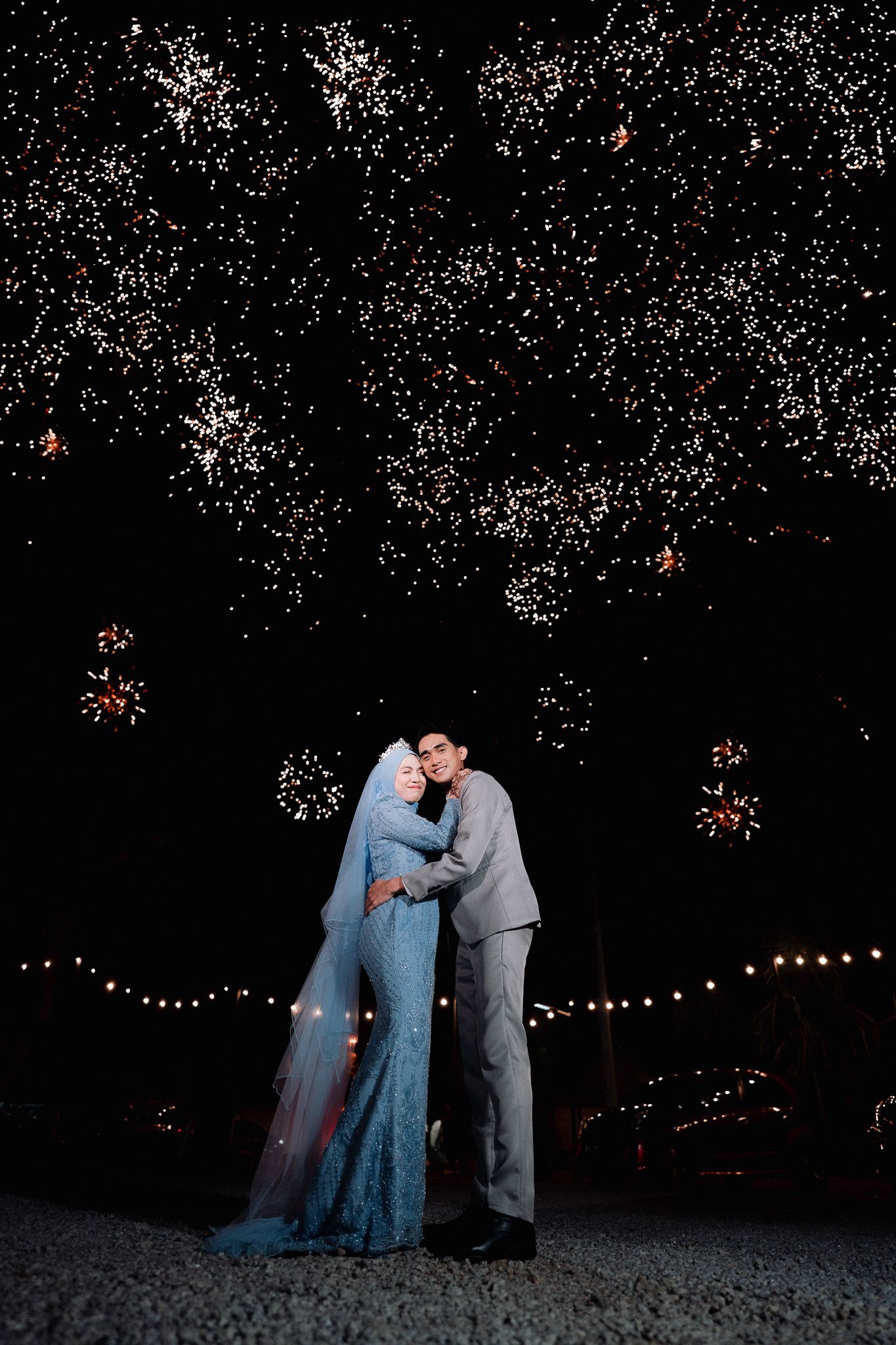 A couple embraces under a stunning fireworks display at night, the bride wearing a blue hijab gown and groom in a gray suit.
