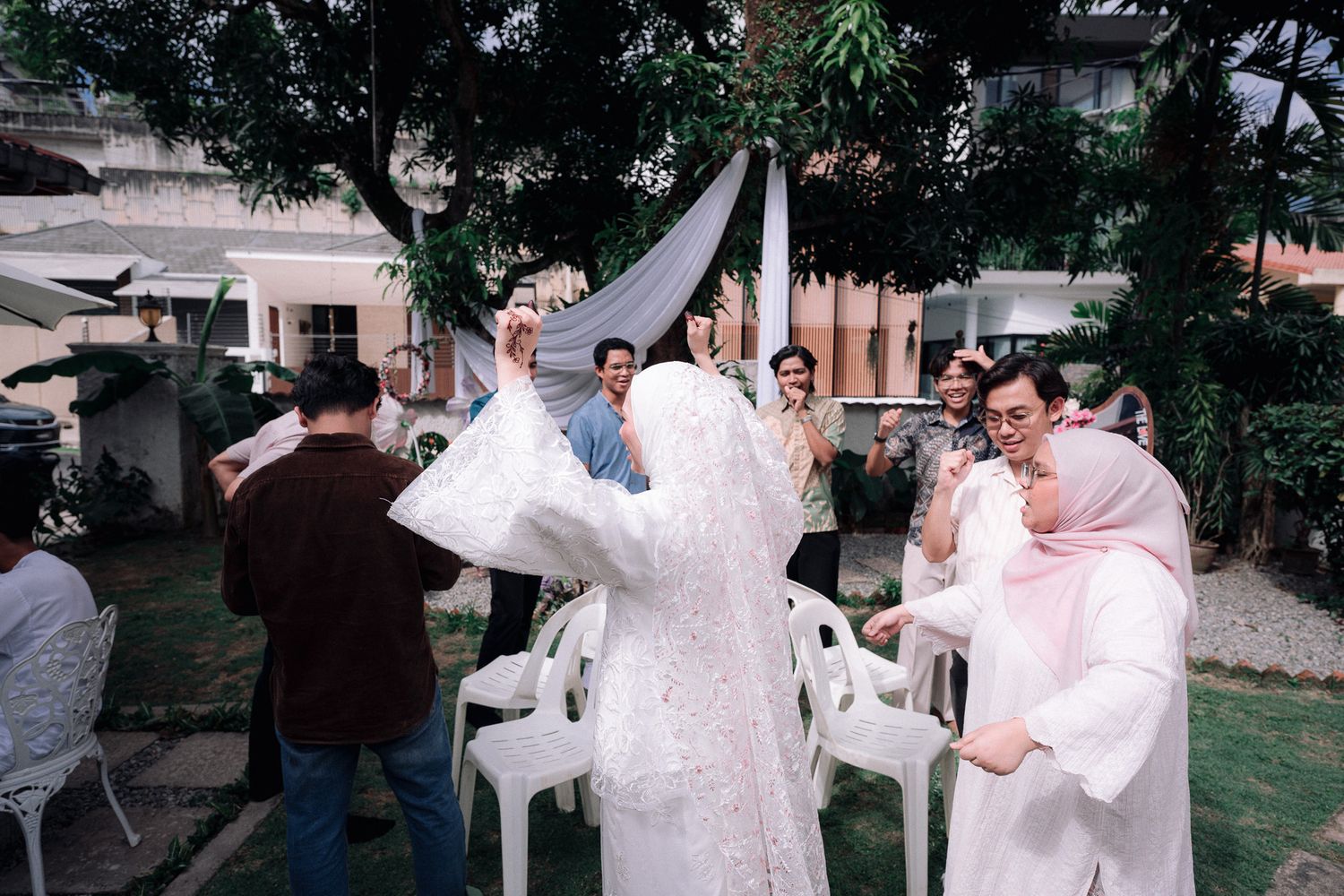Outdoor wedding celebration with guests in white attire gathered under a tree, bride in hijab dancing joyfully.