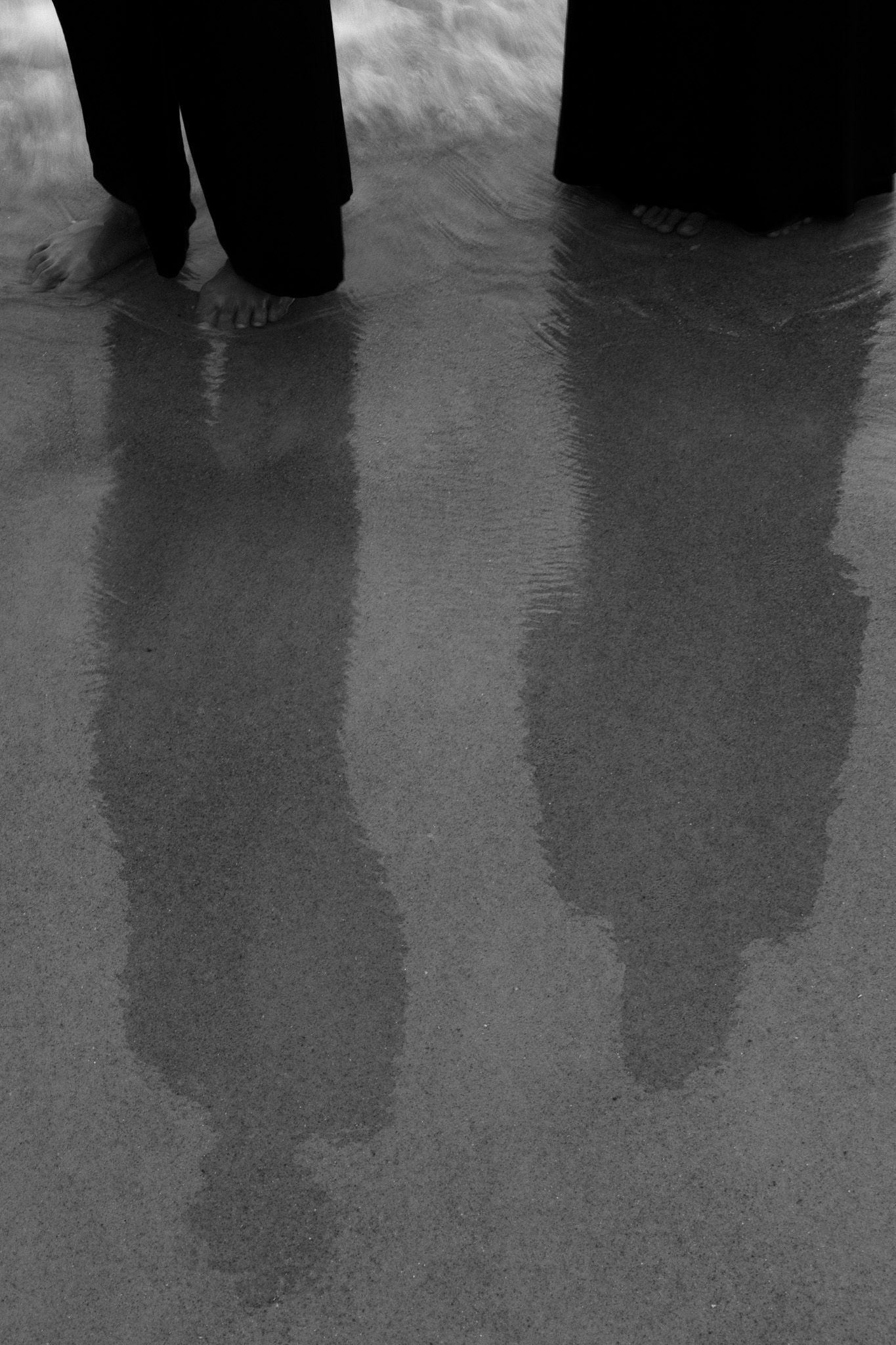 Black and white photo of two people standing barefoot on wet sand with long reflections stretching below them.
