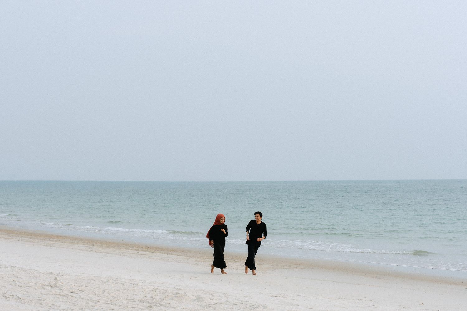Two people in black outfits walking on a serene white sand beach with calm ocean waves and overcast sky.