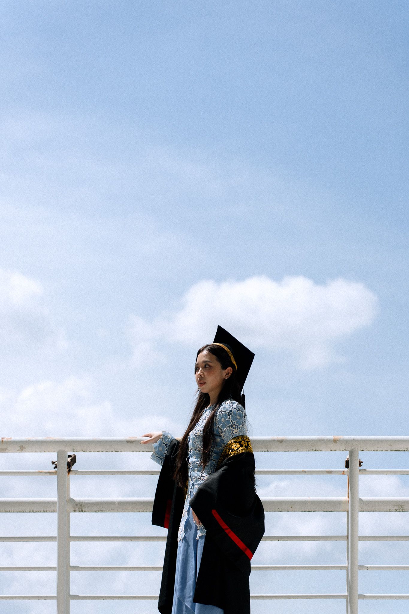 A young woman in graduation cap and gown poses confidently by a railing under a bright blue sky.