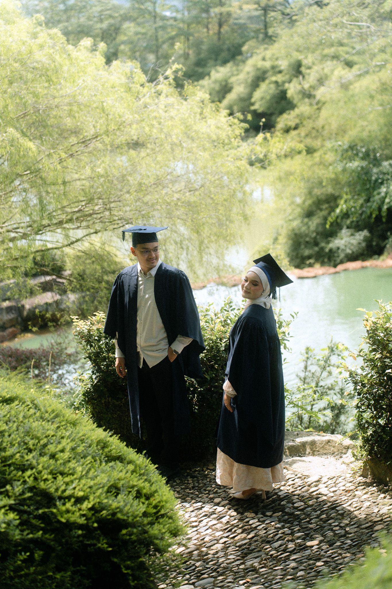 Two graduates in navy blue caps and gowns pose in a lush garden setting near a serene pond.