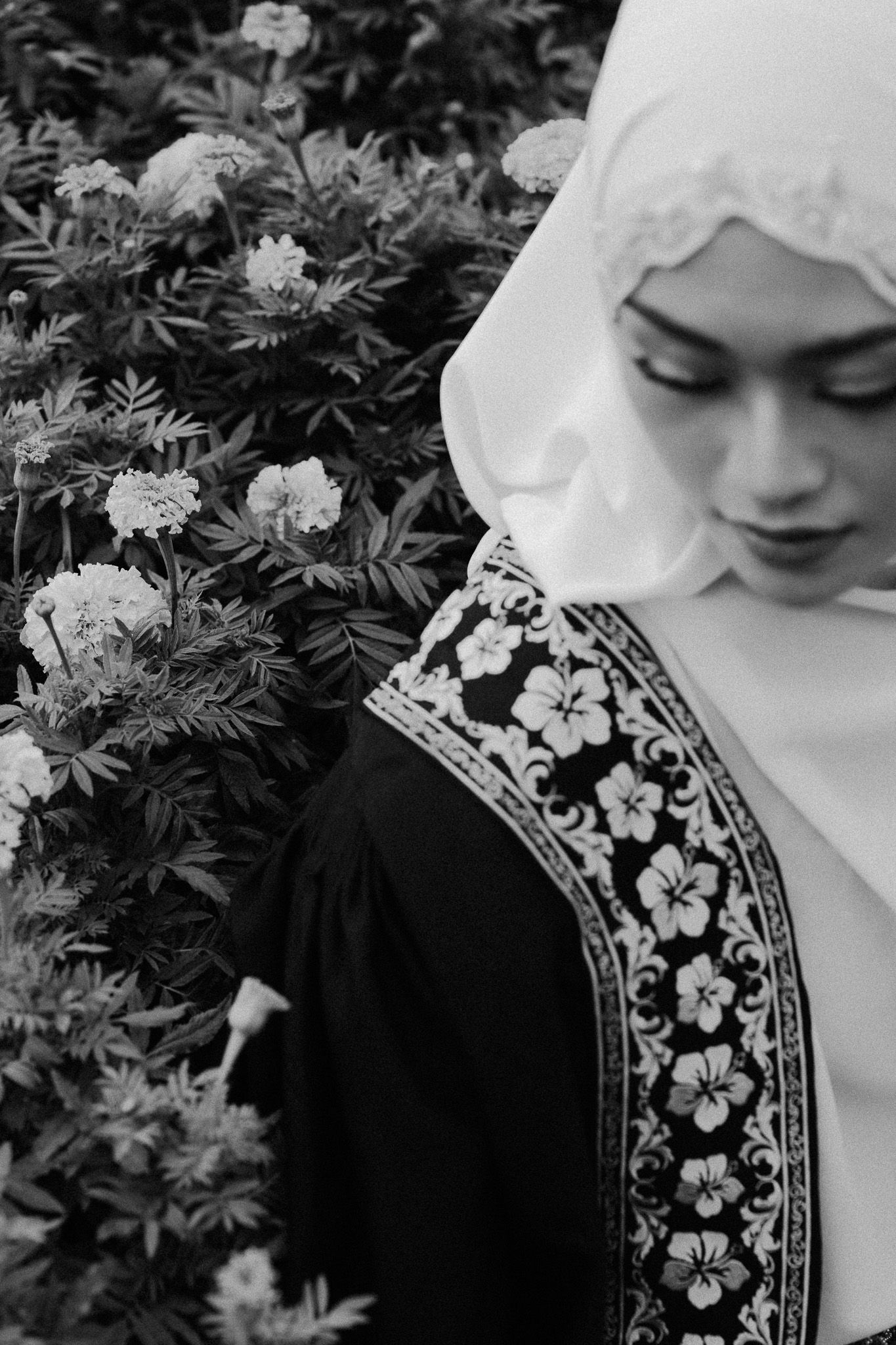 Black and white portrait of a young woman in hijab and floral embroidered dress among blooming flowers.