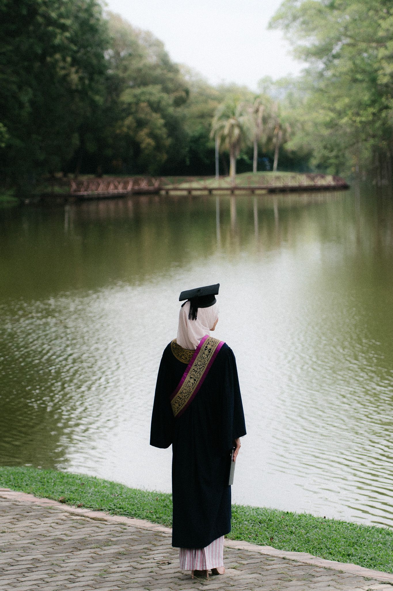 Graduate in black gown and cap stands by a serene lake surrounded by lush green trees.