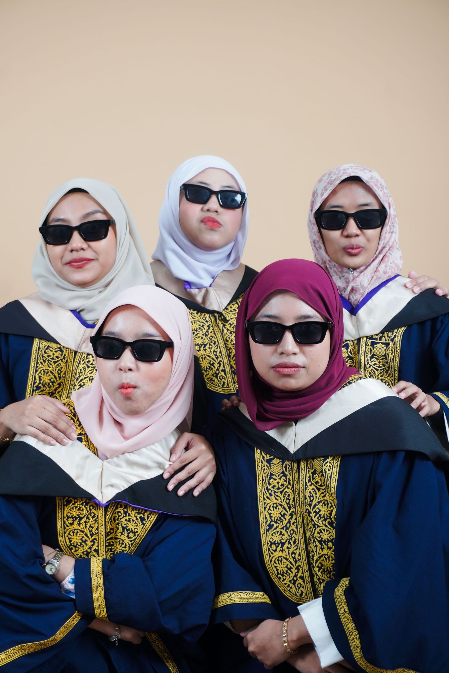 Five women in graduation robes and hijabs wearing sunglasses pose playfully for a group photo.