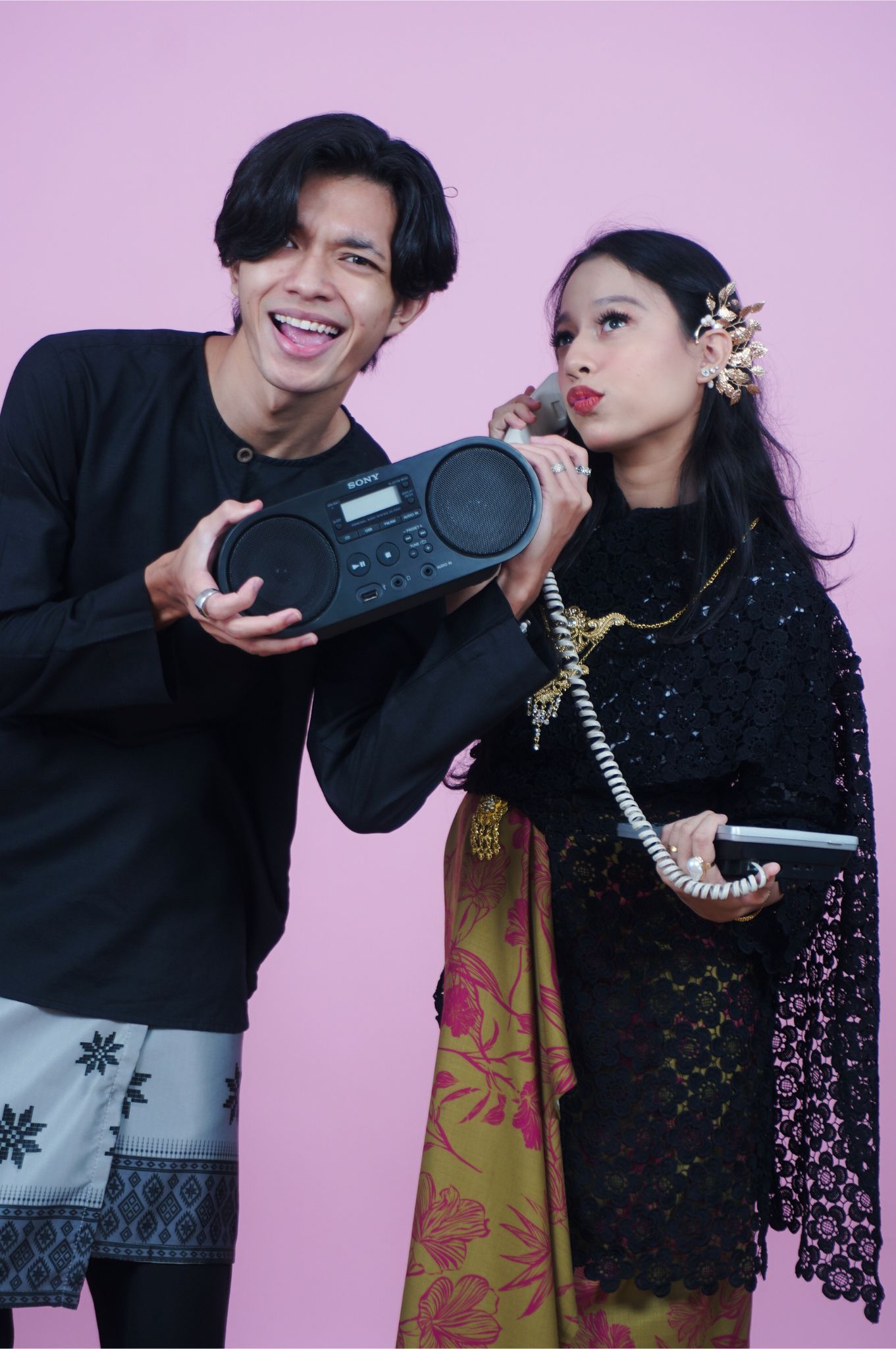A young man holding a Sony boombox and a woman in traditional kebaya attire posing together on a pink background.