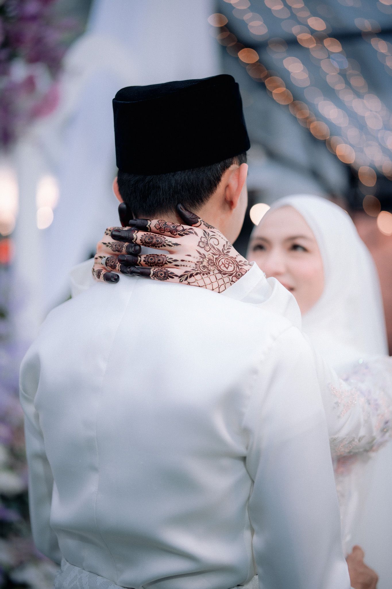 Muslim couple in white wedding attire, groom wearing songkok hat with henna-adorned hands facing bride in hijab.