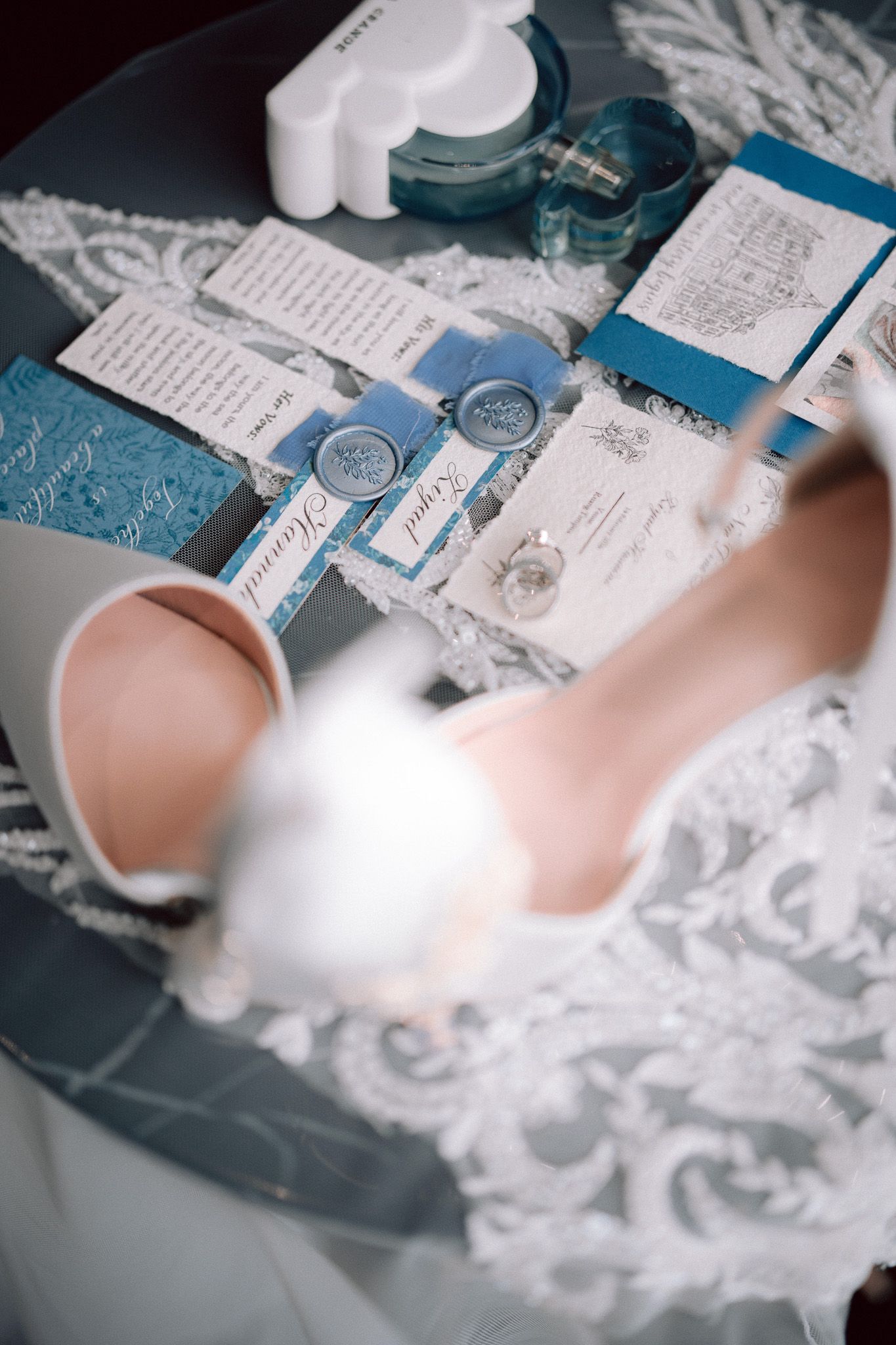 Elegant wedding flat lay with blue velvet invitations, wax seals, bridal heels, and perfume on a gray table.