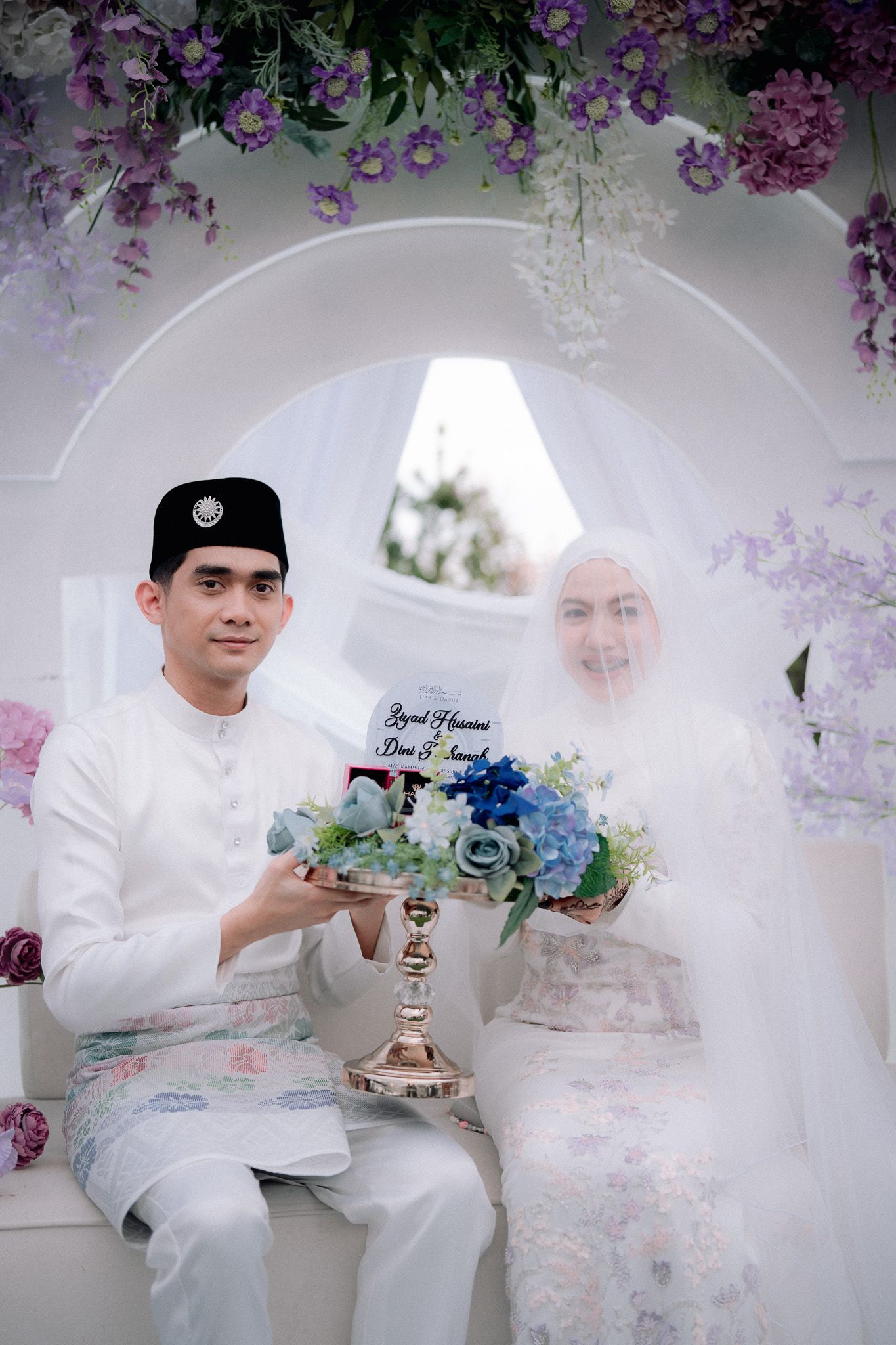 Malay couple in traditional white wedding attire seated under floral arch holding decorative arrangement.