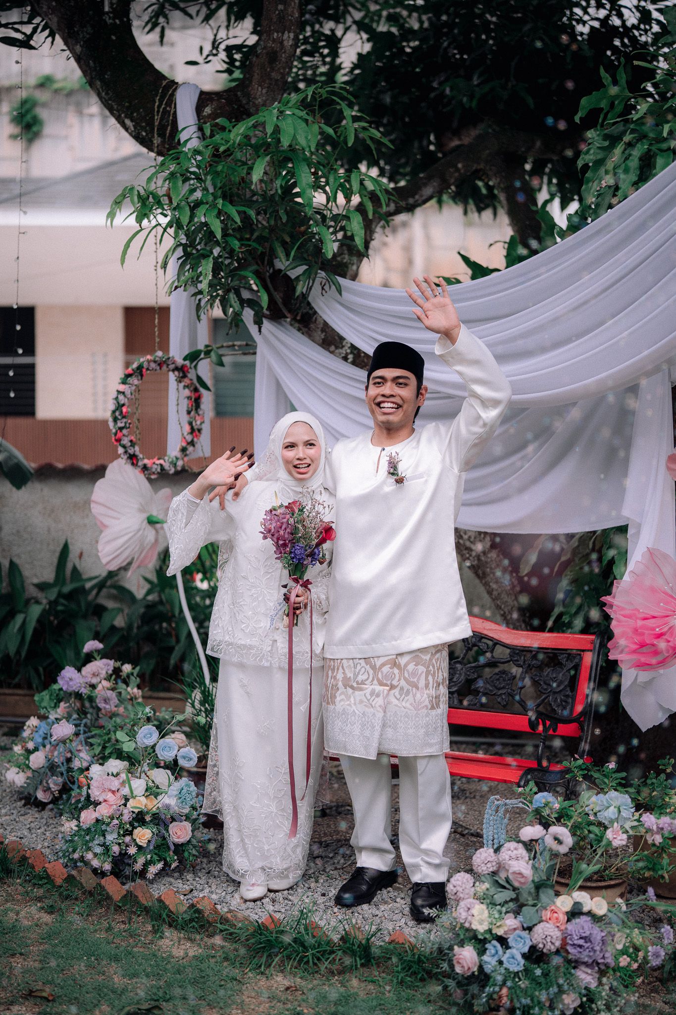 A joyful couple in traditional white Malay wedding attire waves and smiles outdoors, surrounded by colorful floral decorations.