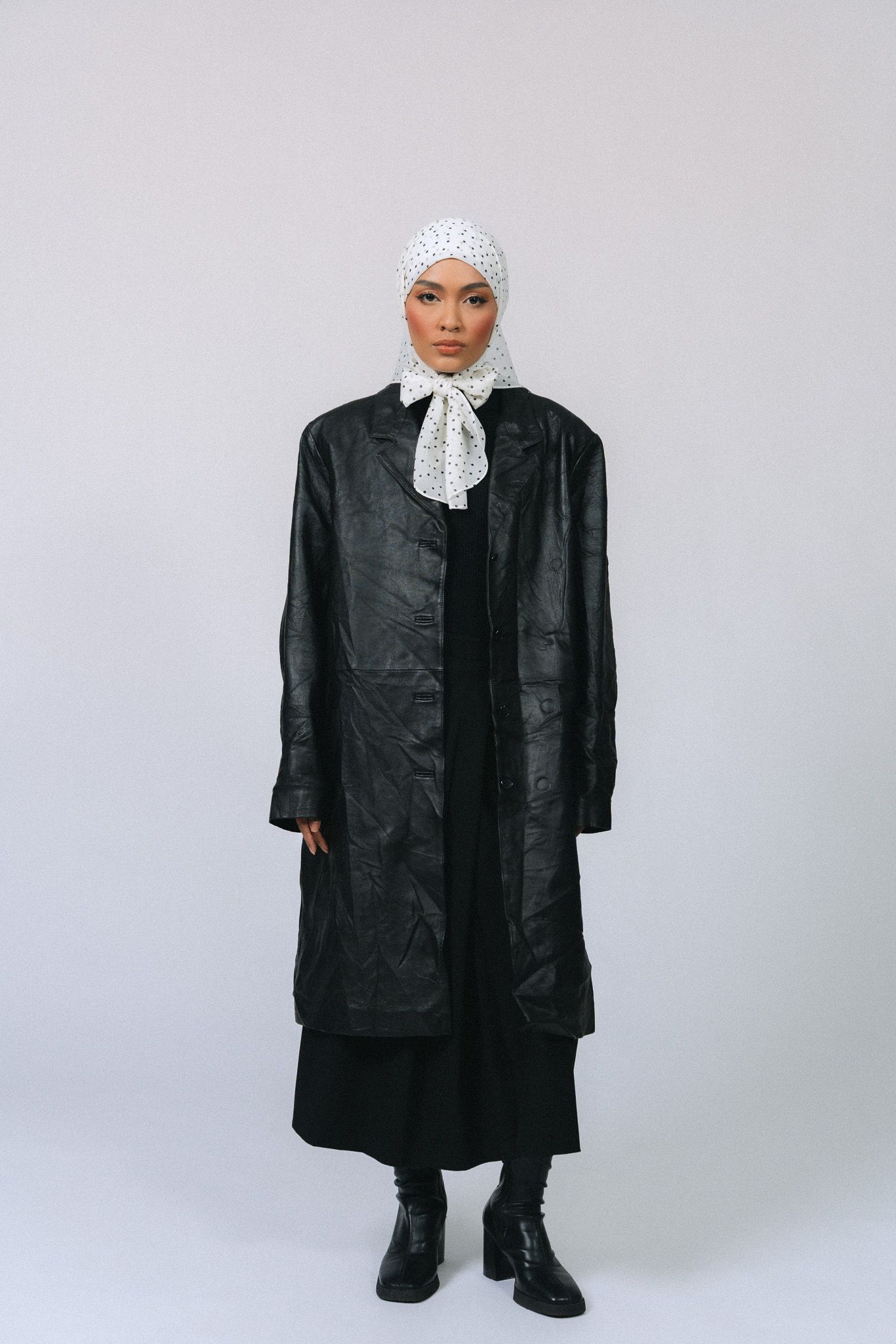 Woman in black leather coat and white polka dot hijab with bow tie, standing against gray background.