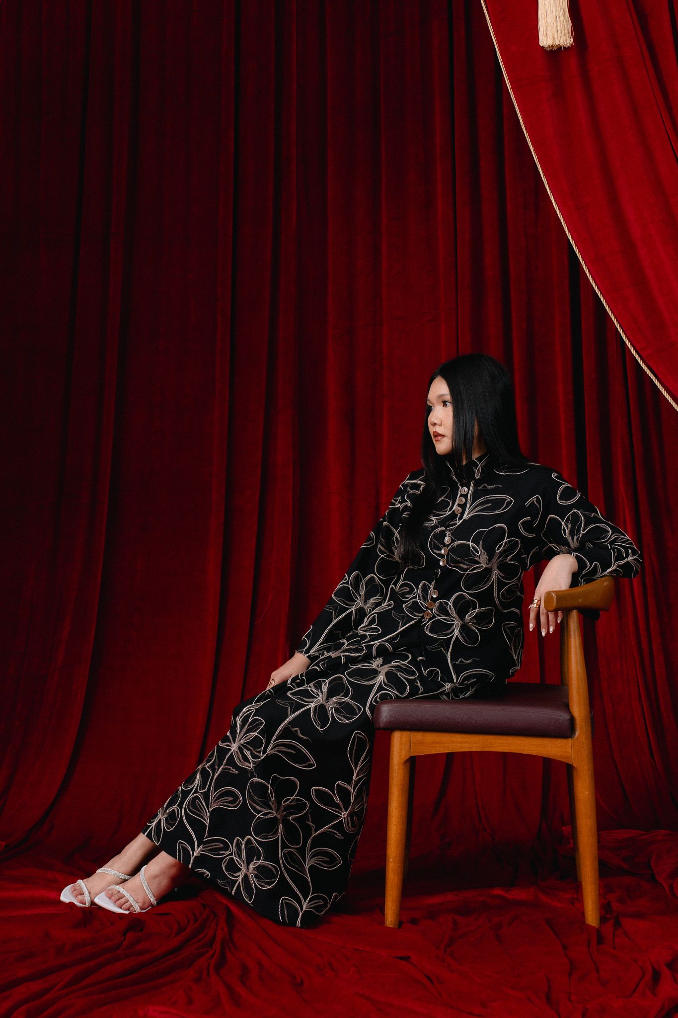 Woman in black floral dress seated on wooden chair against red velvet curtain backdrop.