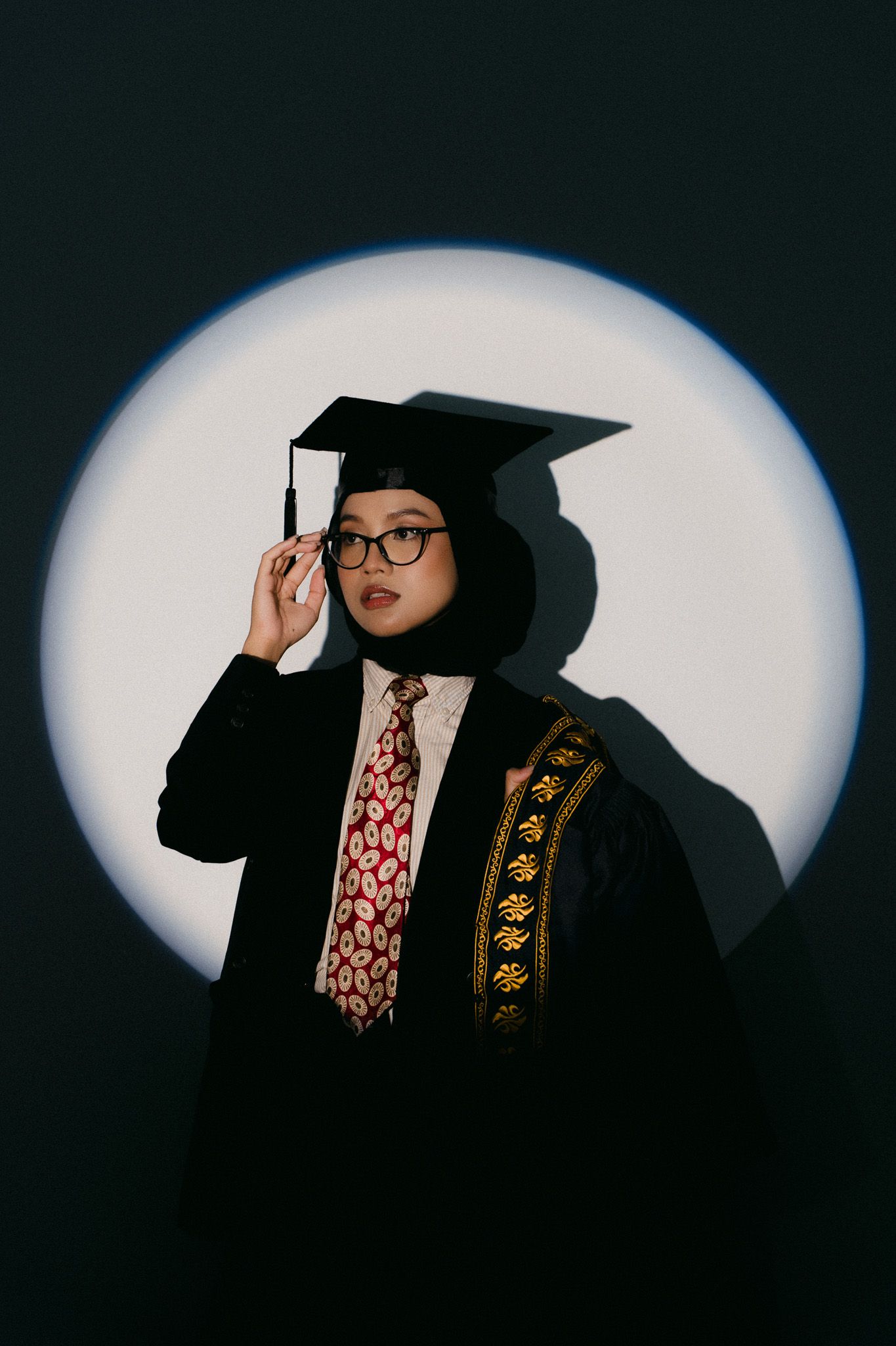 Graduate in black cap, gown, hijab, and glasses poses confidently under spotlight against dark background.