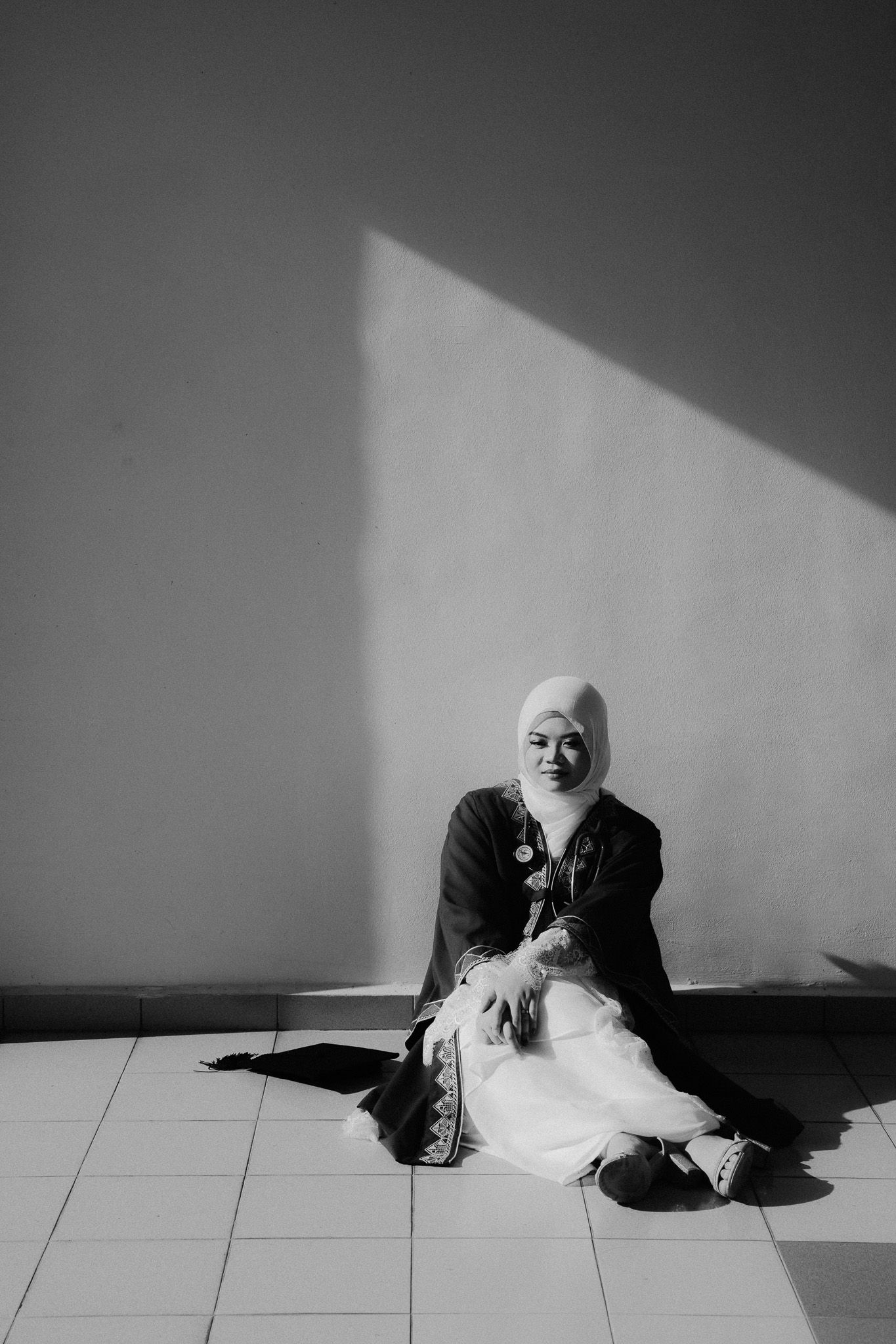Black and white photo of a woman in hijab sitting gracefully on tiled floor against sunlit wall.