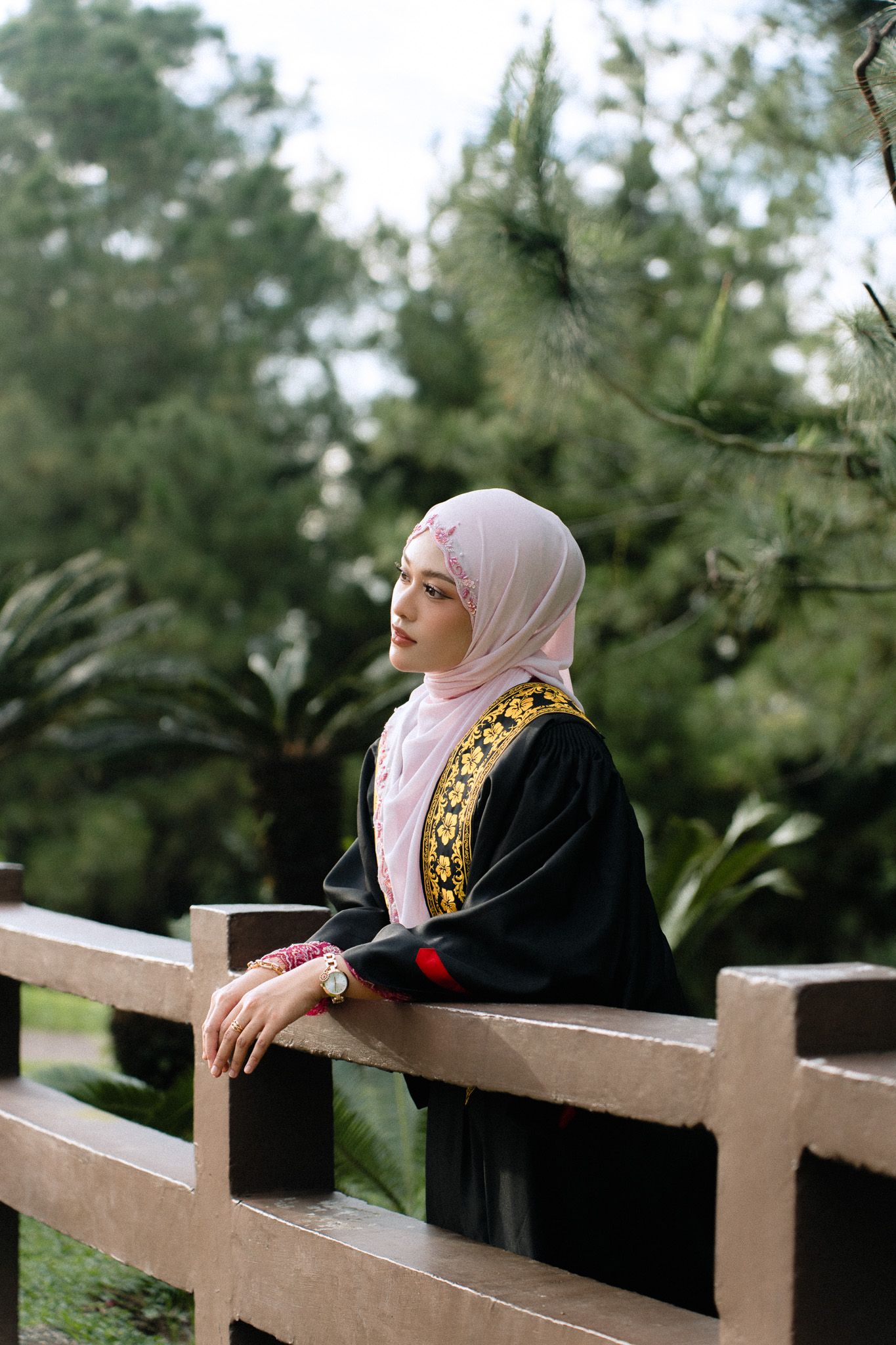 Young Muslim woman in black abaya and pink hijab leaning on wooden fence in lush green park.