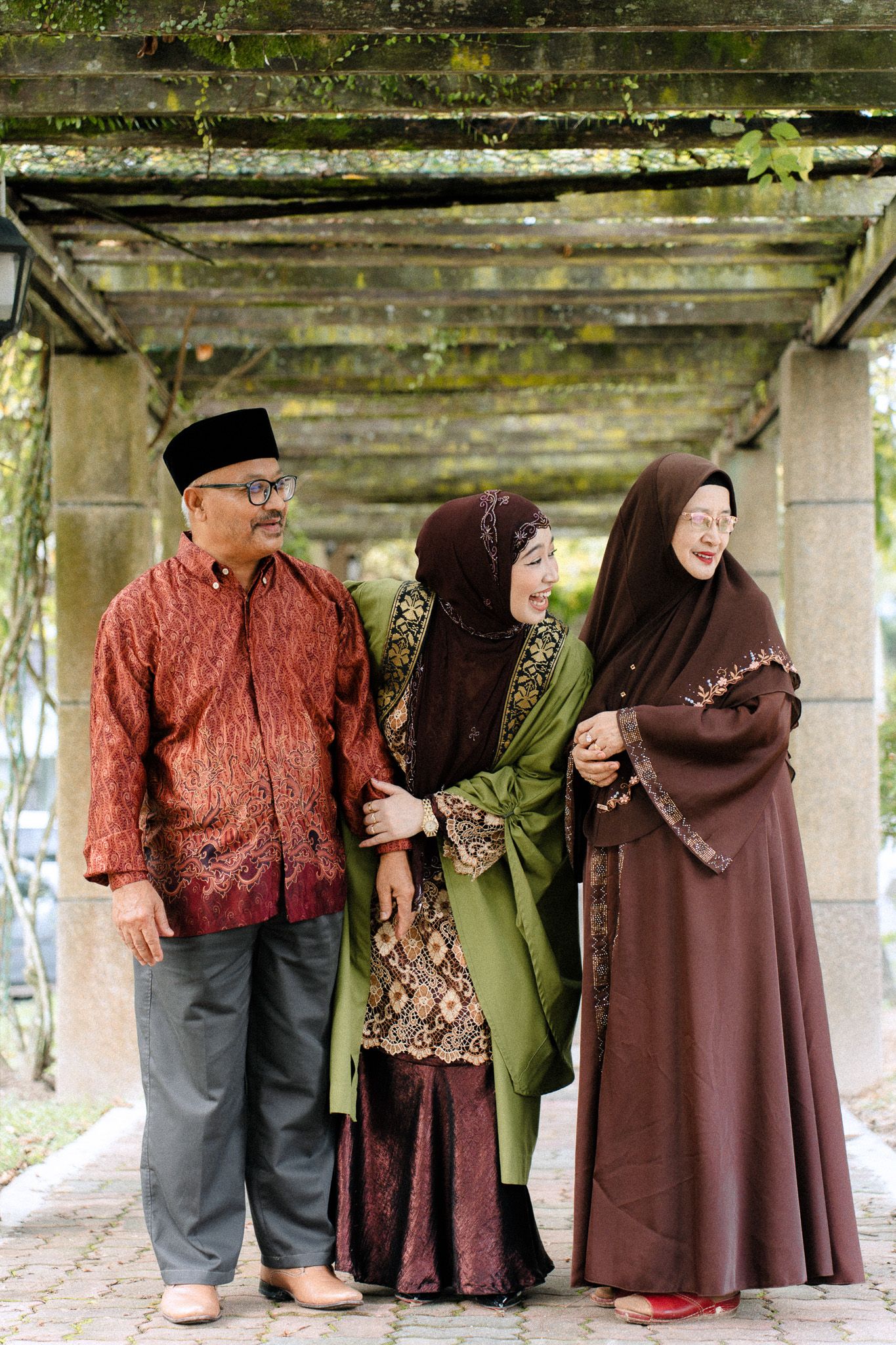 Multi-generational Muslim family wearing traditional Indonesian batik and hijab standing together under a wooden pergola.