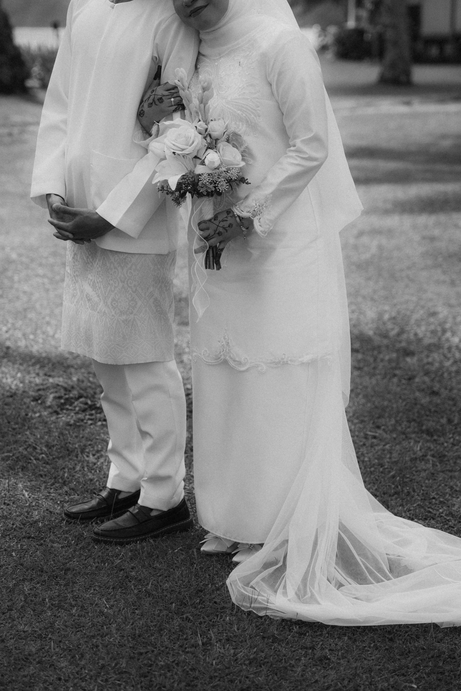 Black and white wedding photo showing close-up of couple in white attire holding bouquet and embracing outdoors.