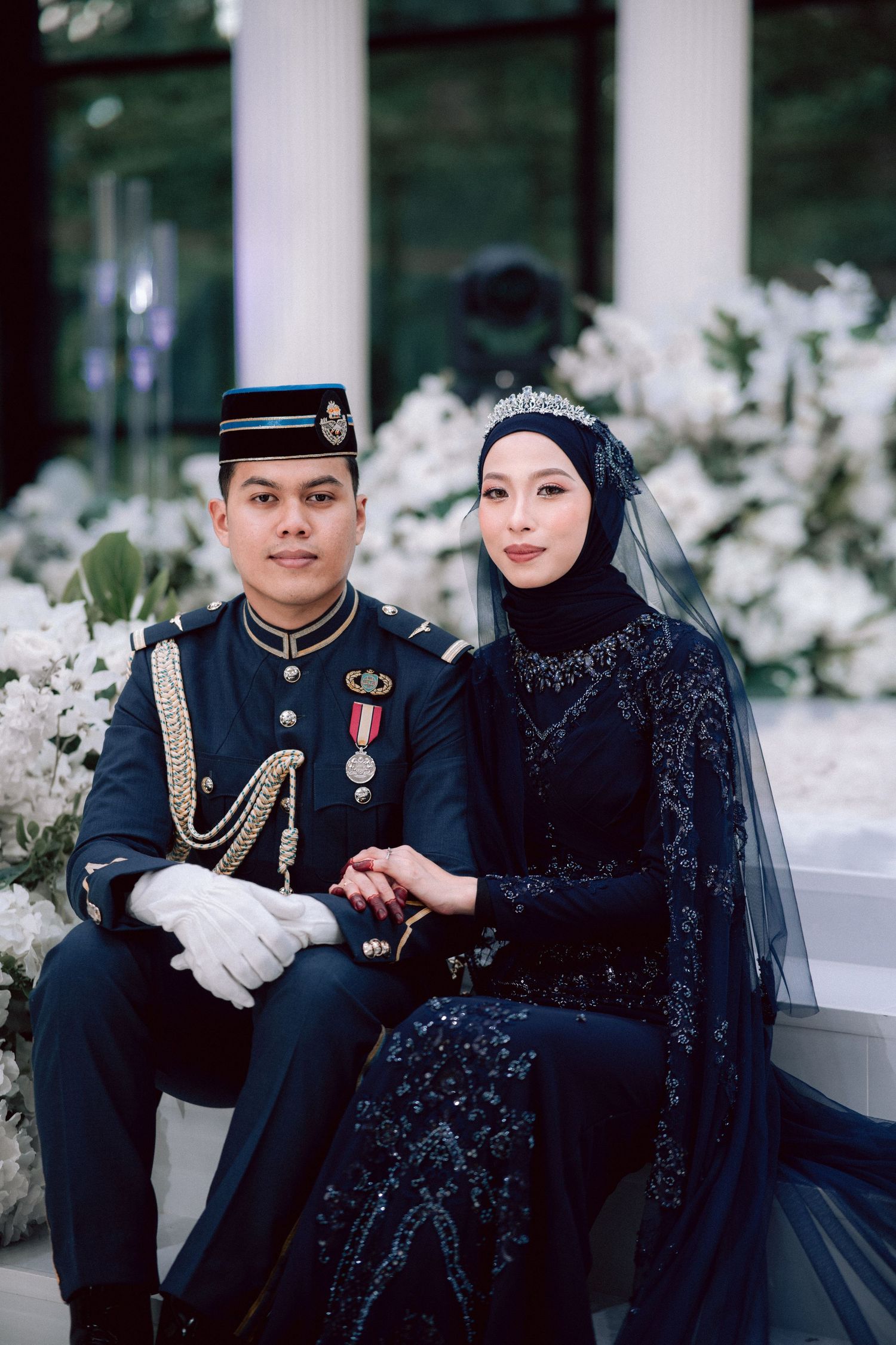 Military wedding photo featuring elegant navy blue dress uniform and black beaded formal gown with hijab against white florals.