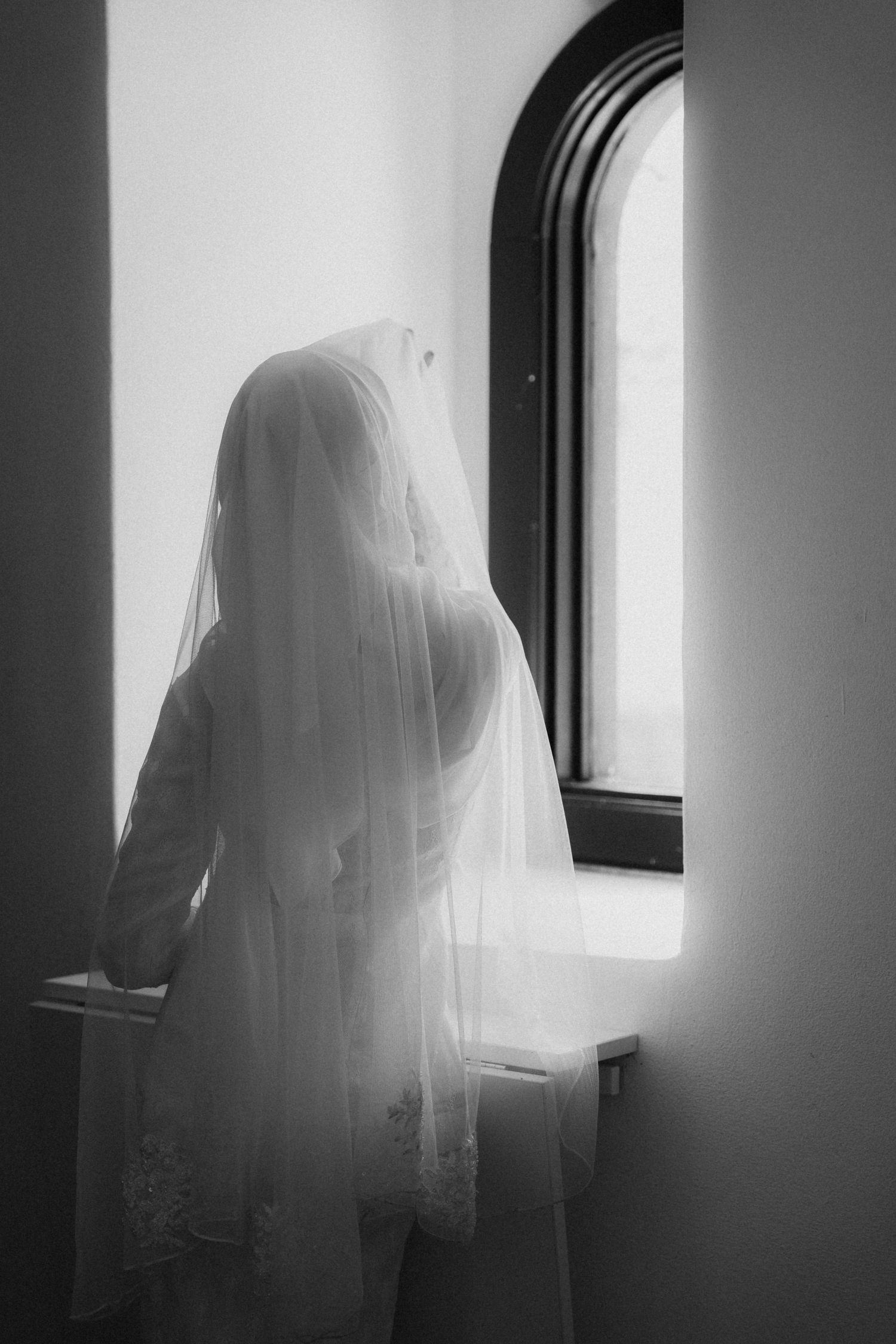 A silhouetted figure in a white dress stands by an arched window in a black and white photograph.