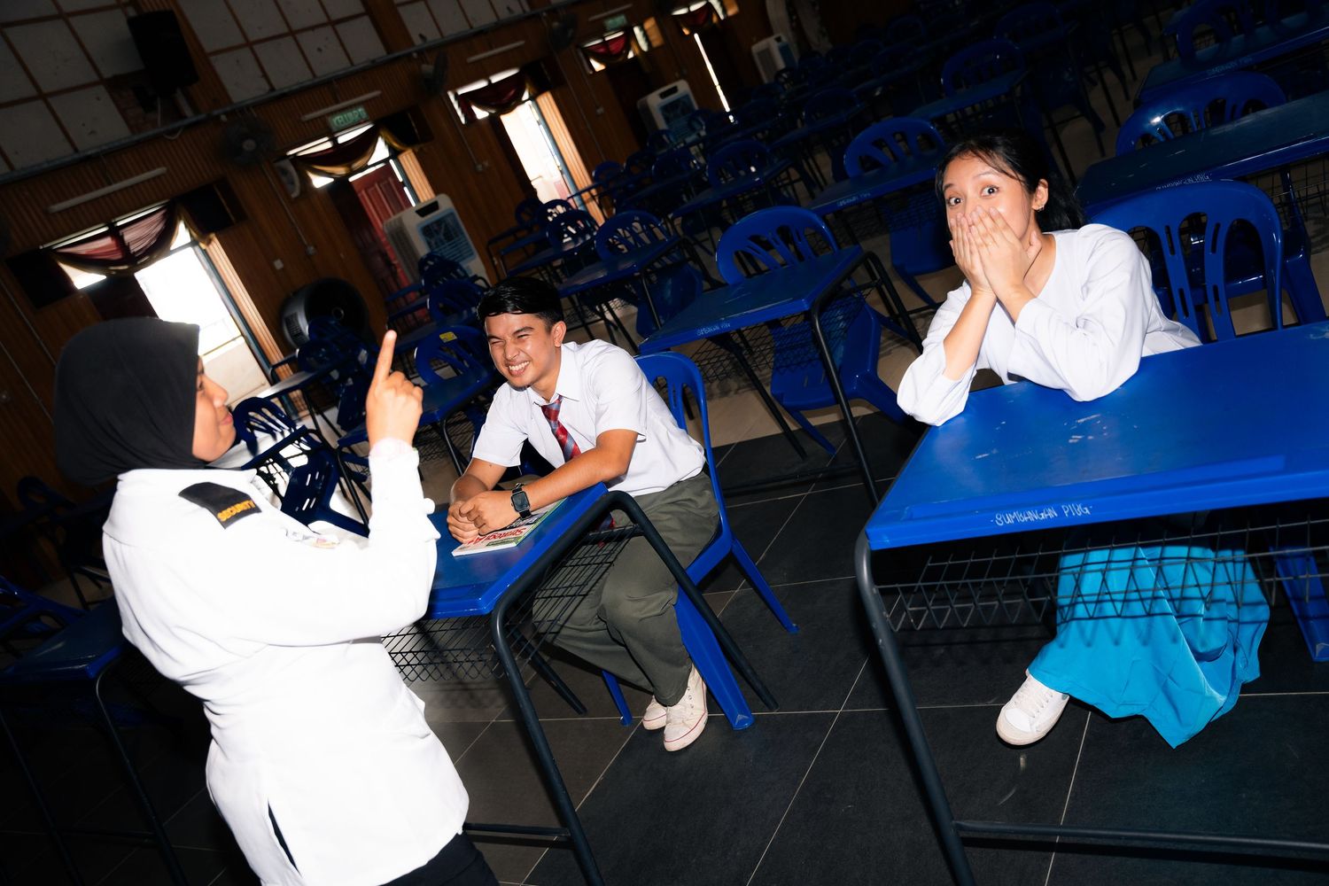 Students sit on blue chairs in a dimly lit space while someone takes photos with a flash camera.