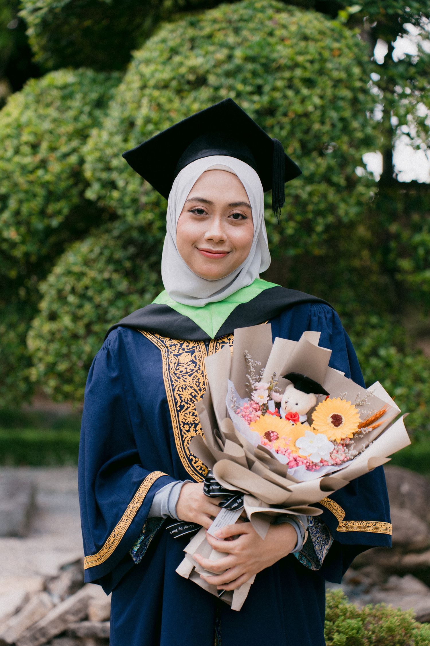 A graduate in traditional academic attire holds a bouquet of flowers and donuts during a celebration outdoors.