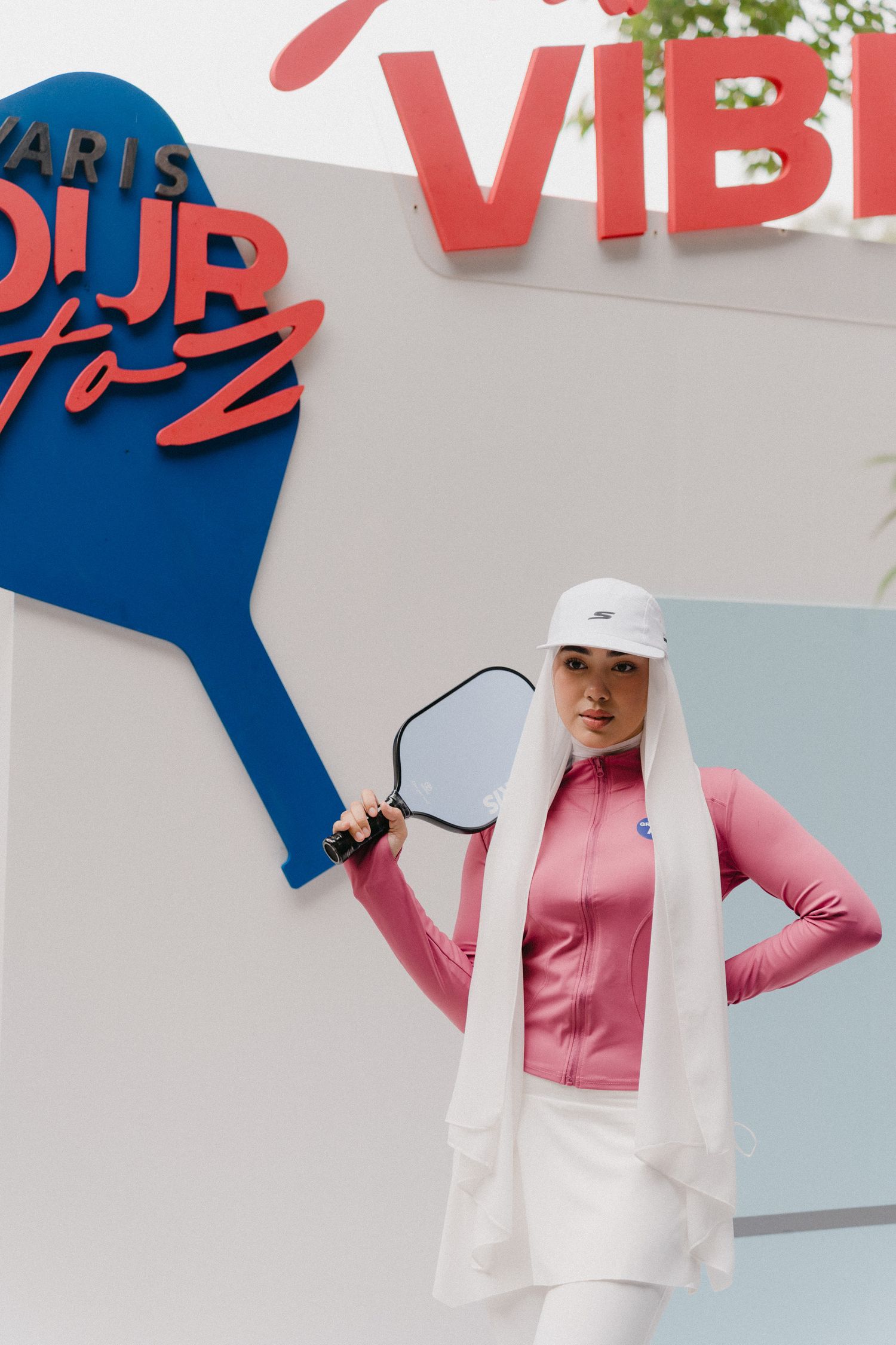 Woman in pink turtleneck and white baseball cap poses against white wall with blue geometric shapes.
