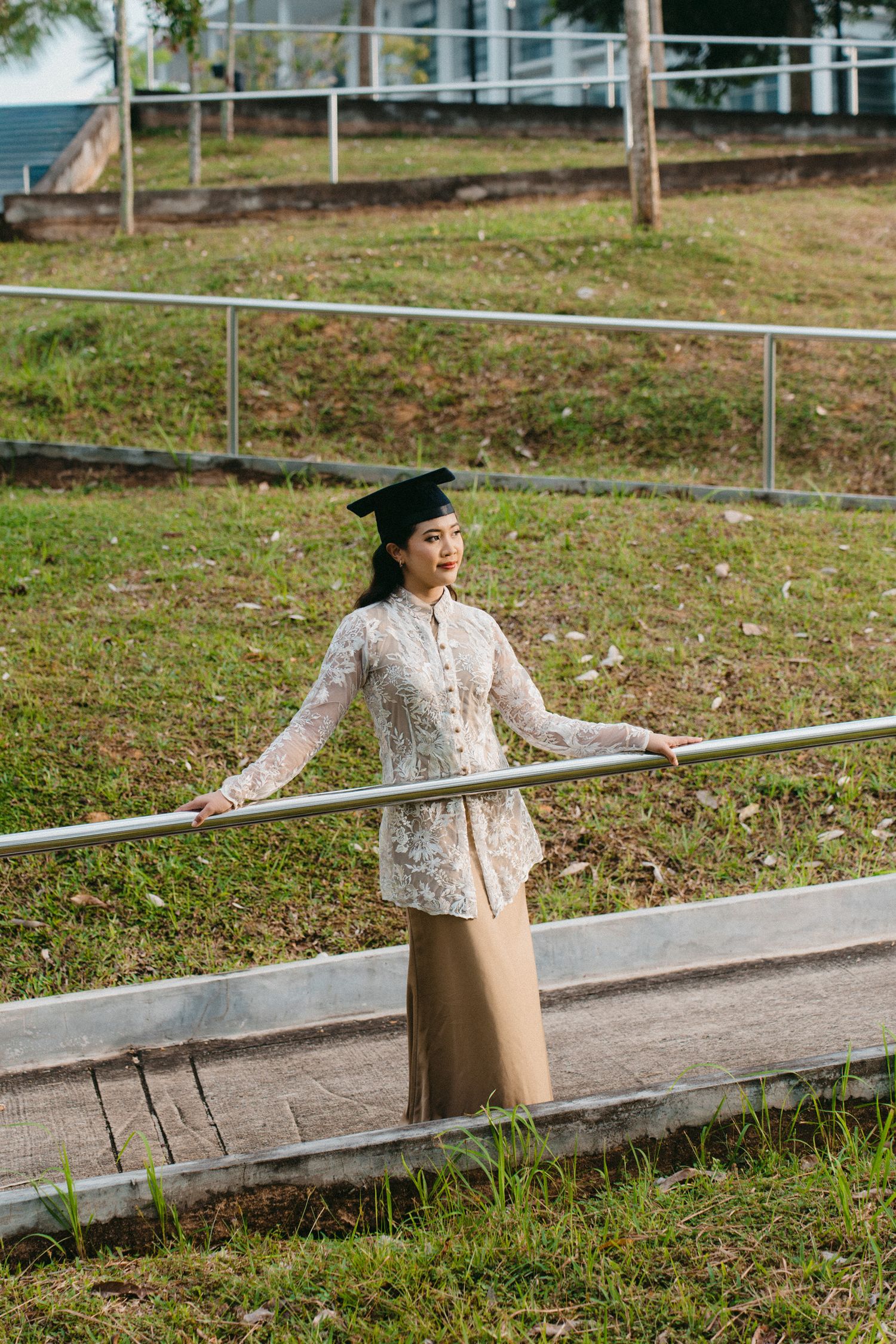 Graduate in a floral print top poses casually on a metal railing outdoors on a grassy slope.