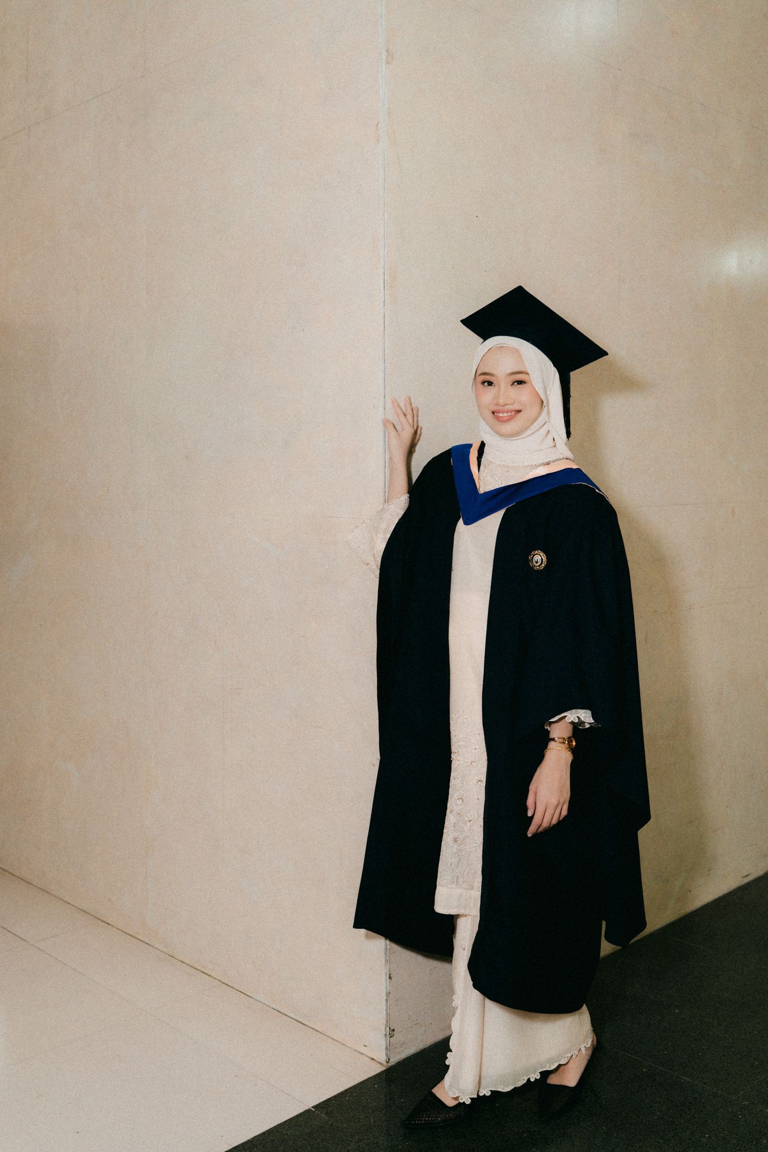 Graduate in traditional black academic regalia and white dress poses against beige wall during commencement ceremony.