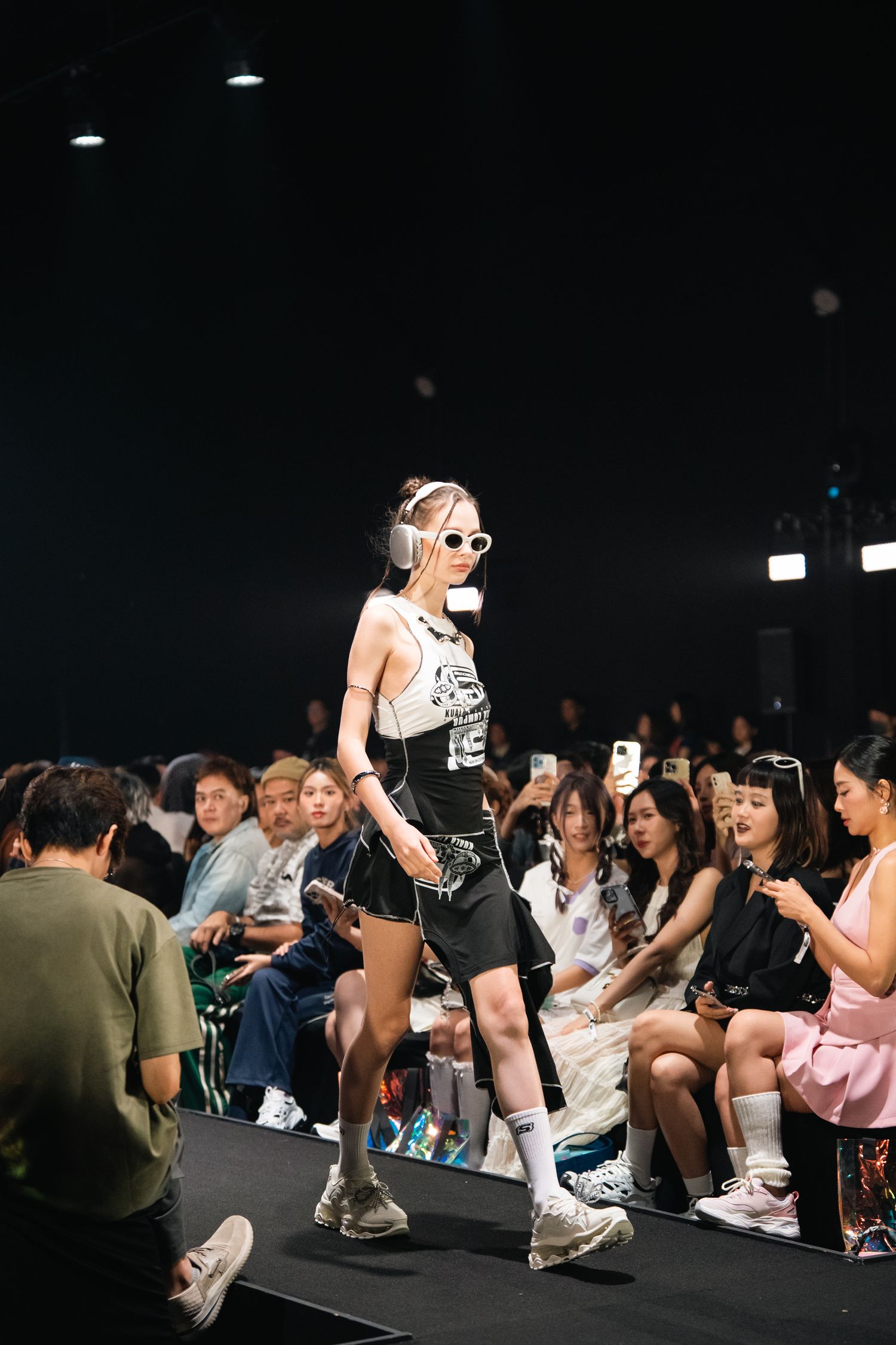 A model walks the runway in athletic wear while audience members take photos on their phones.