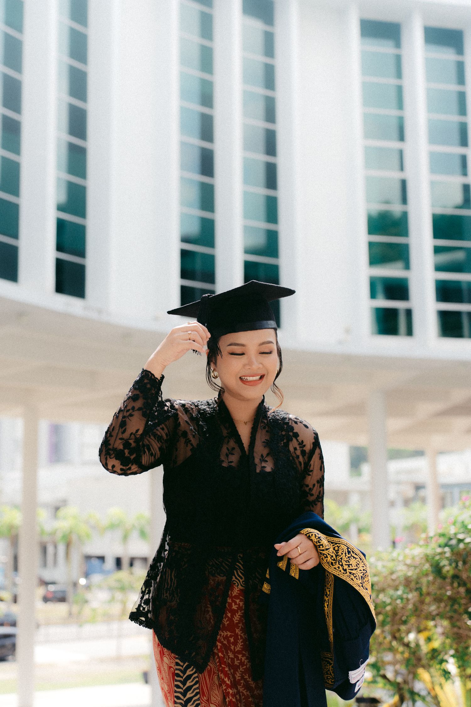 Graduate wearing black lace top and traditional batik adjusts mortarboard cap while smiling outdoors by modern building.