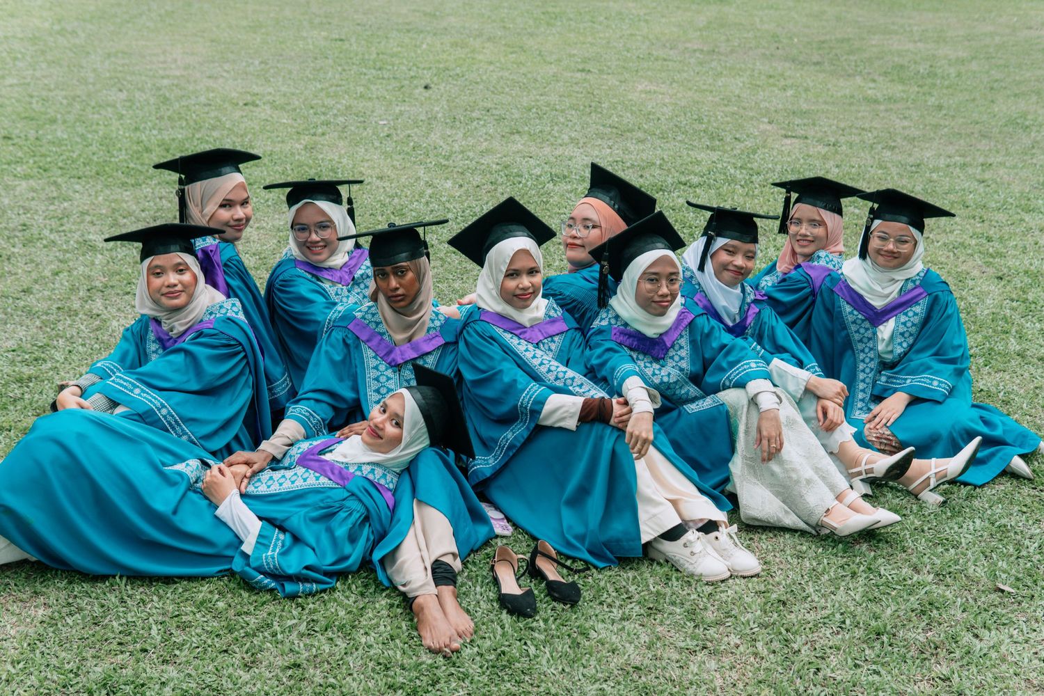 Group of graduates in turquoise robes lounging on grass during graduation photo session.