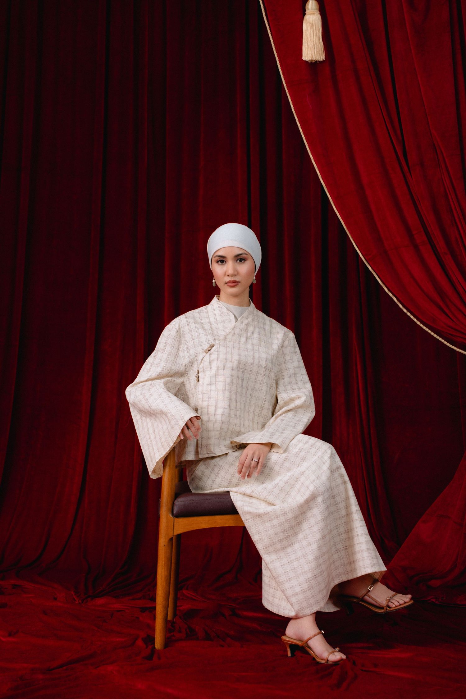 Figure in a white checkered dress and headpiece poses dramatically against deep red velvet curtains in a studio setting.