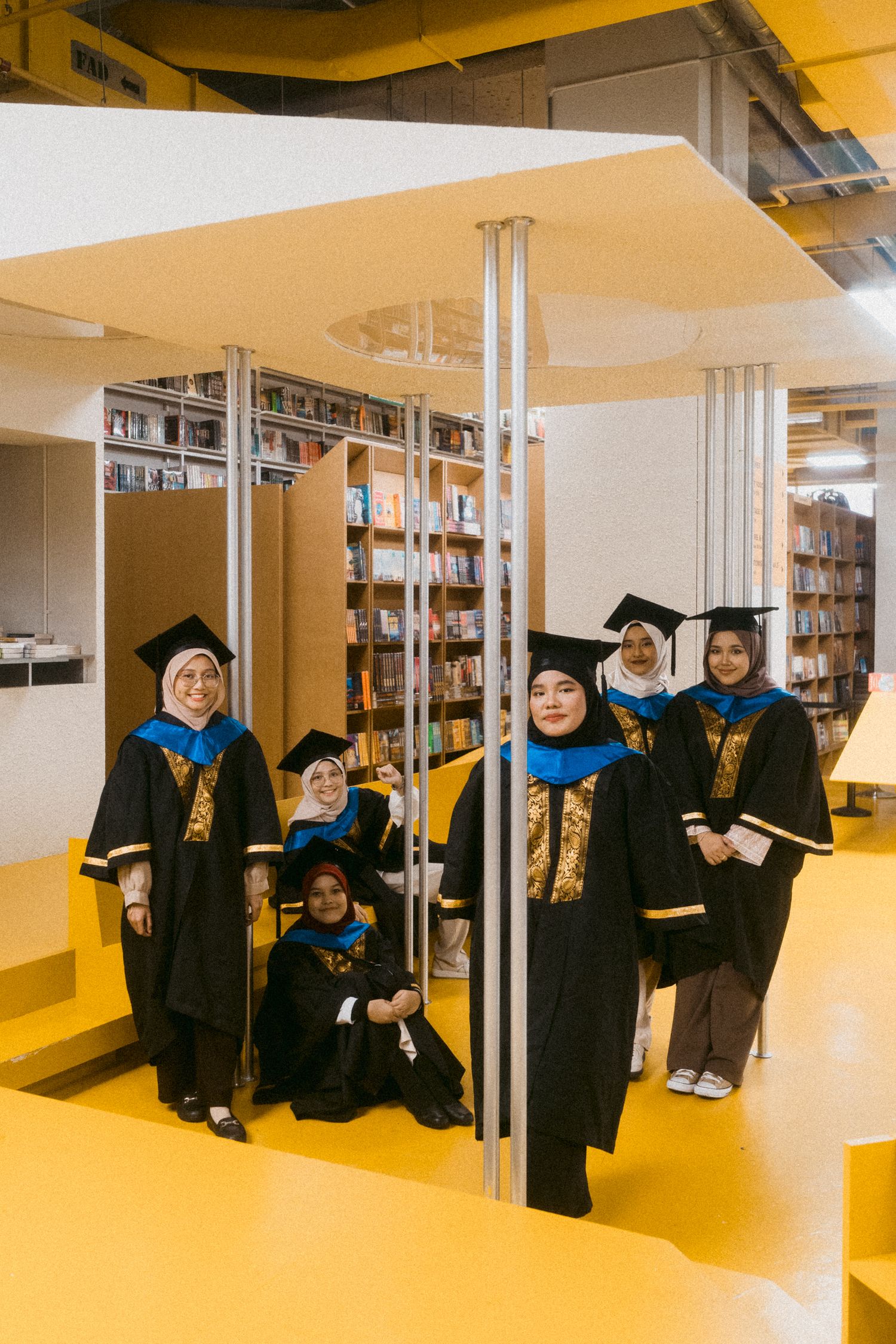 Graduates in black robes with blue stoles gather between library shelves.