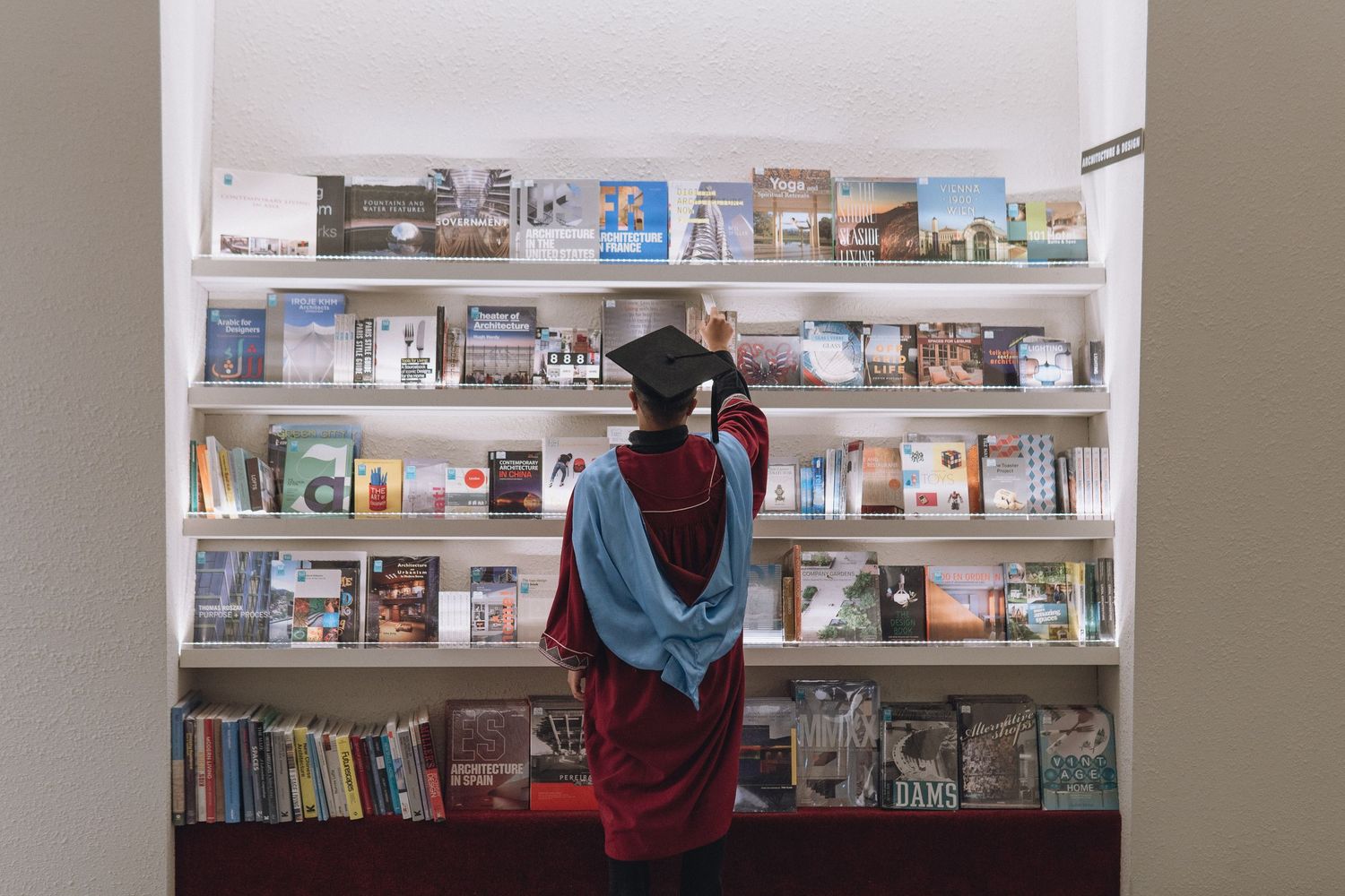 Graduate in maroon and blue regalia browses books on library shelves.