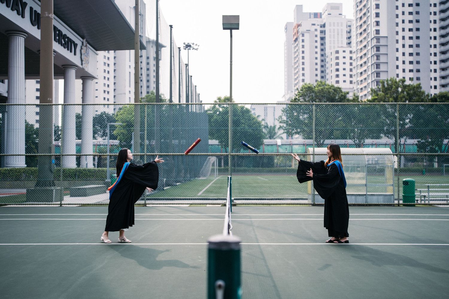 Graduates in academic regalia pose playfully on opposite sides of a tennis court at a university sports facility.
