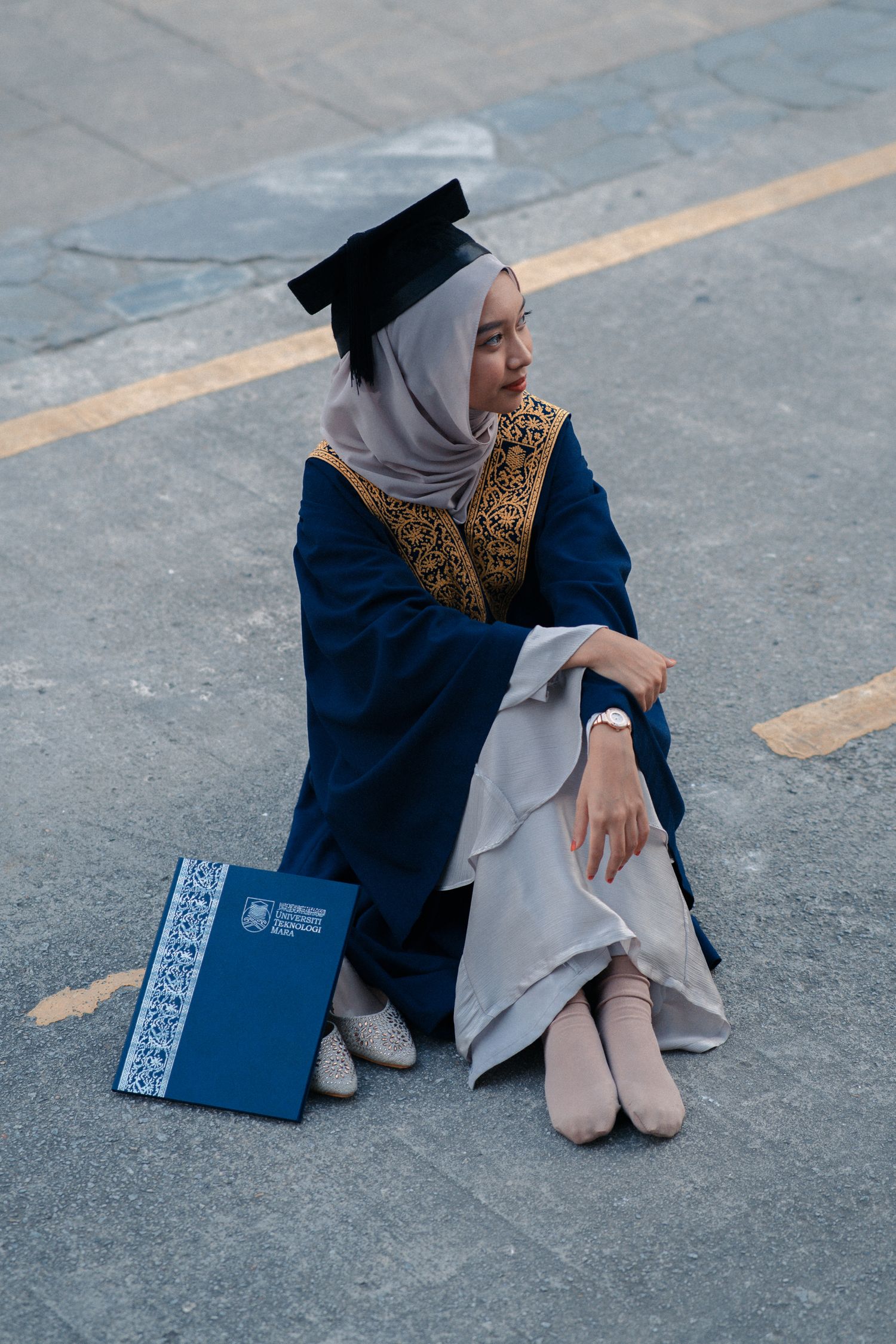 Graduate in traditional dress sitting on pavement with diploma book.