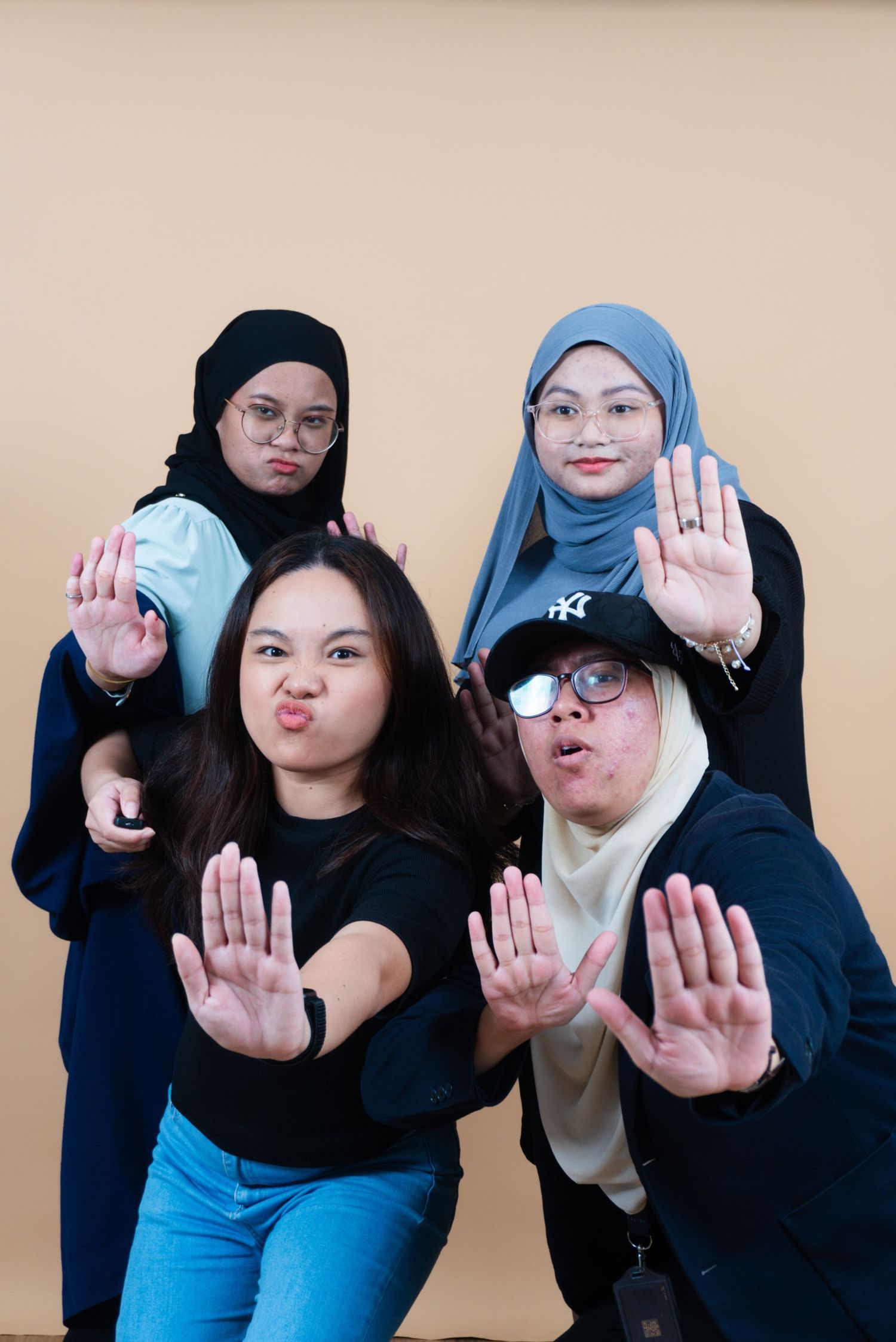 Four young women in hijabs pose playfully, extending hands toward camera against a beige background.
