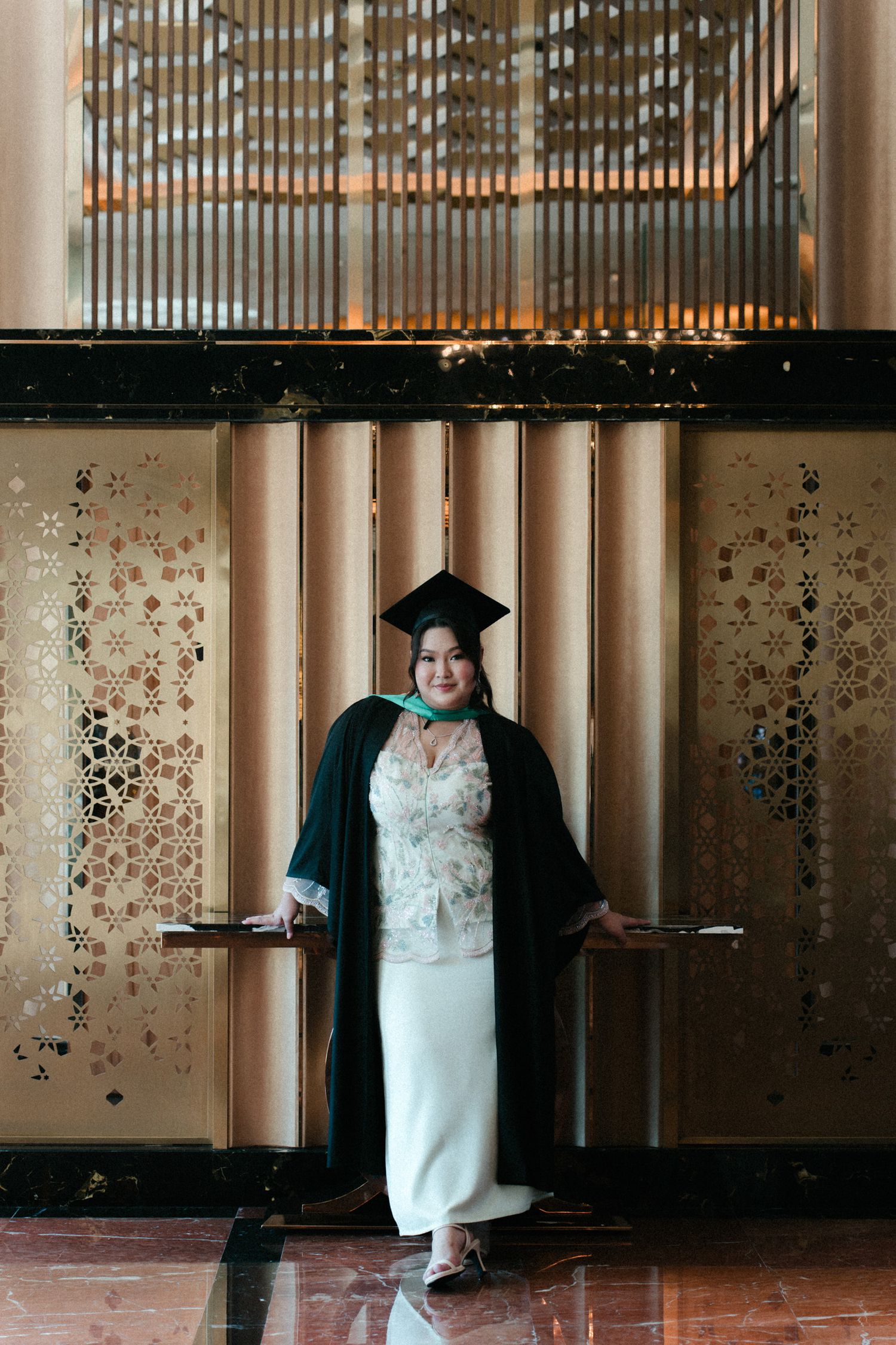 Graduate in black cap and gown stands in front of wall with decorative circular pattern during graduation photoshoot.