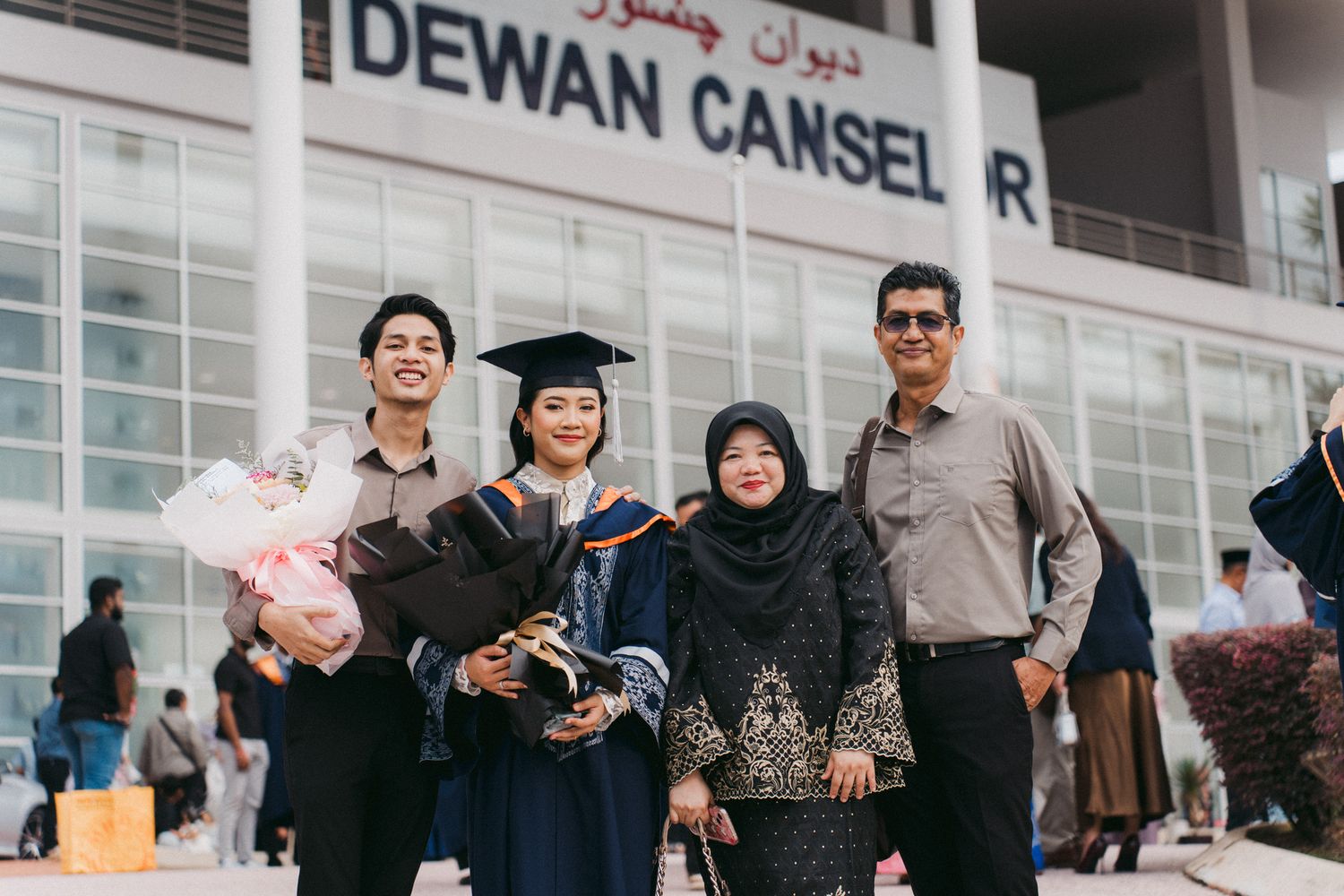 A group of people gather outside a building with a sign that reads 'DEWAN' visible in the background.