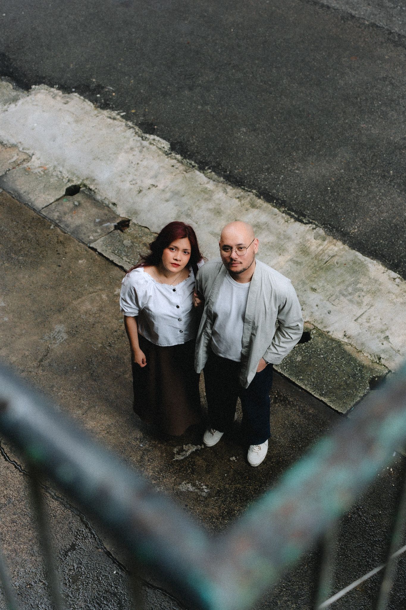 Aerial view of a bald man and woman standing together on a urban sidewalk, looking up at the camera.