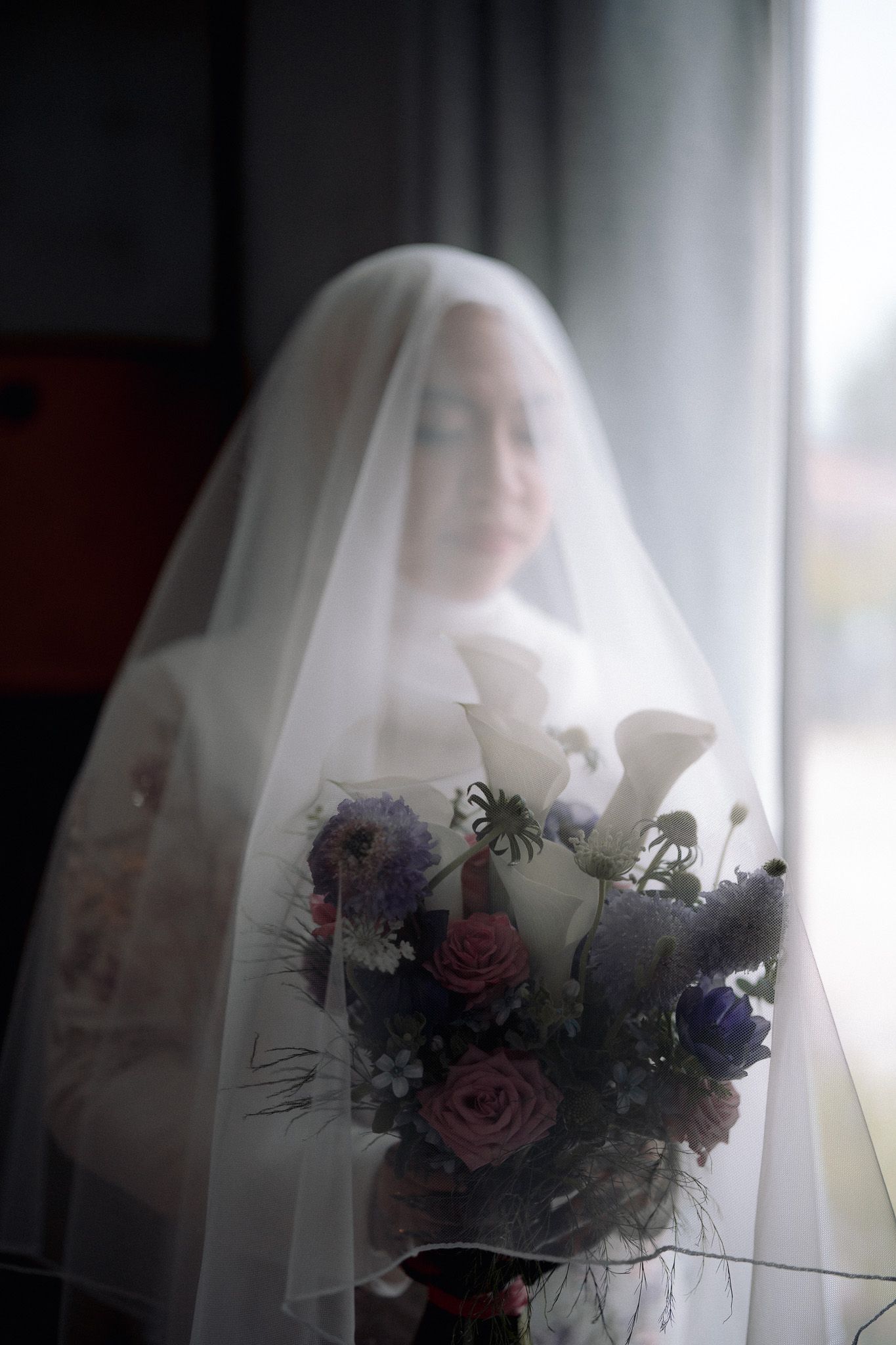 Bride in white veil holding a bouquet of purple and pink flowers near a window on her wedding day.