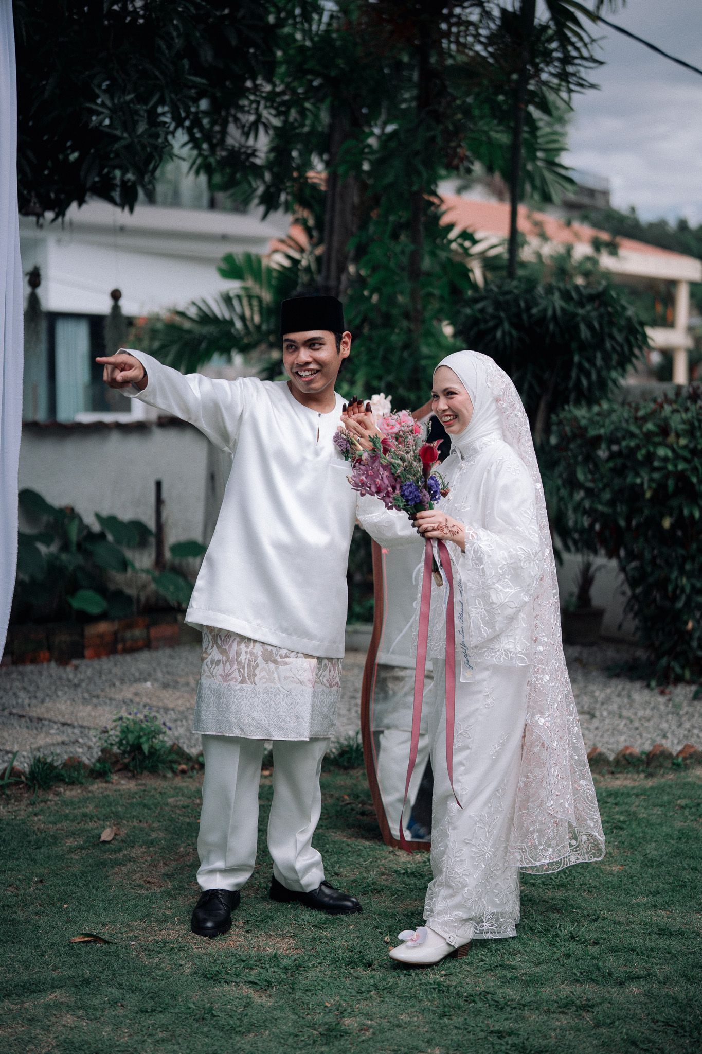 Muslim couple in white traditional attire posing outdoors, groom pointing while bride holds colorful bouquet with pink ribbons.
