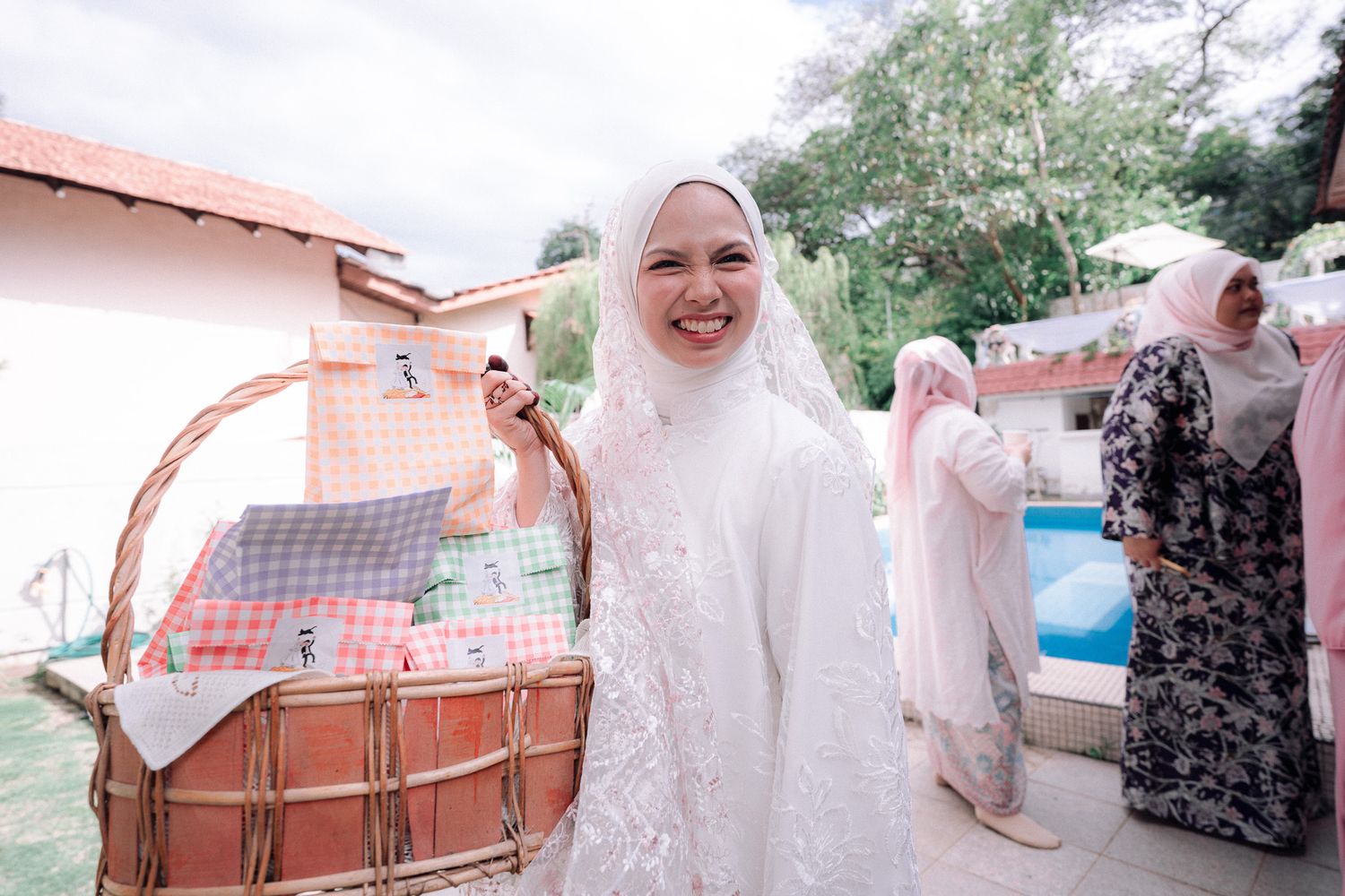 A smiling woman in white hijab and lace dress holds a decorative basket at an outdoor Islamic celebration.